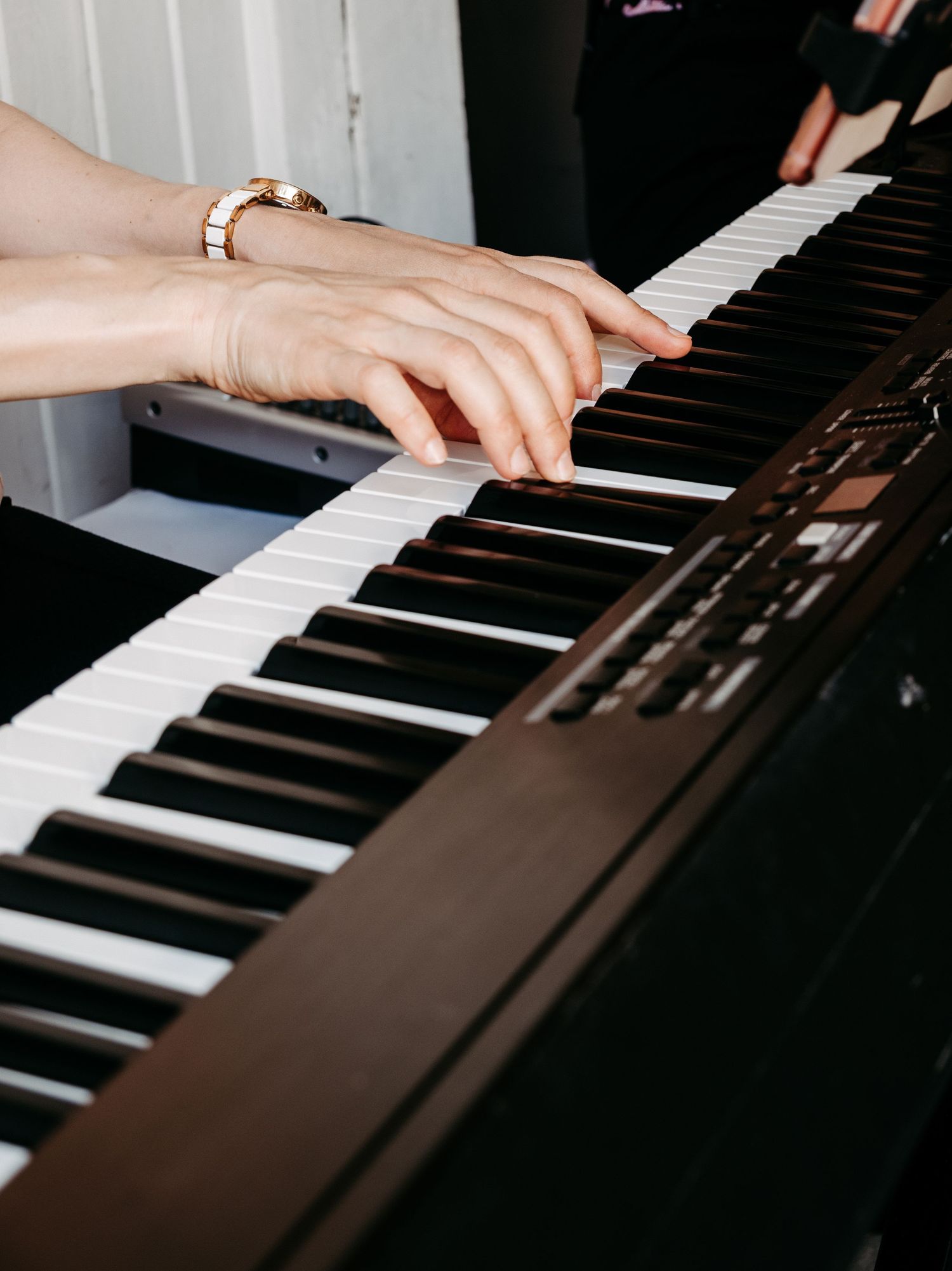Close-up of hands with gold watch playing keys on a dark digital piano keyboard.