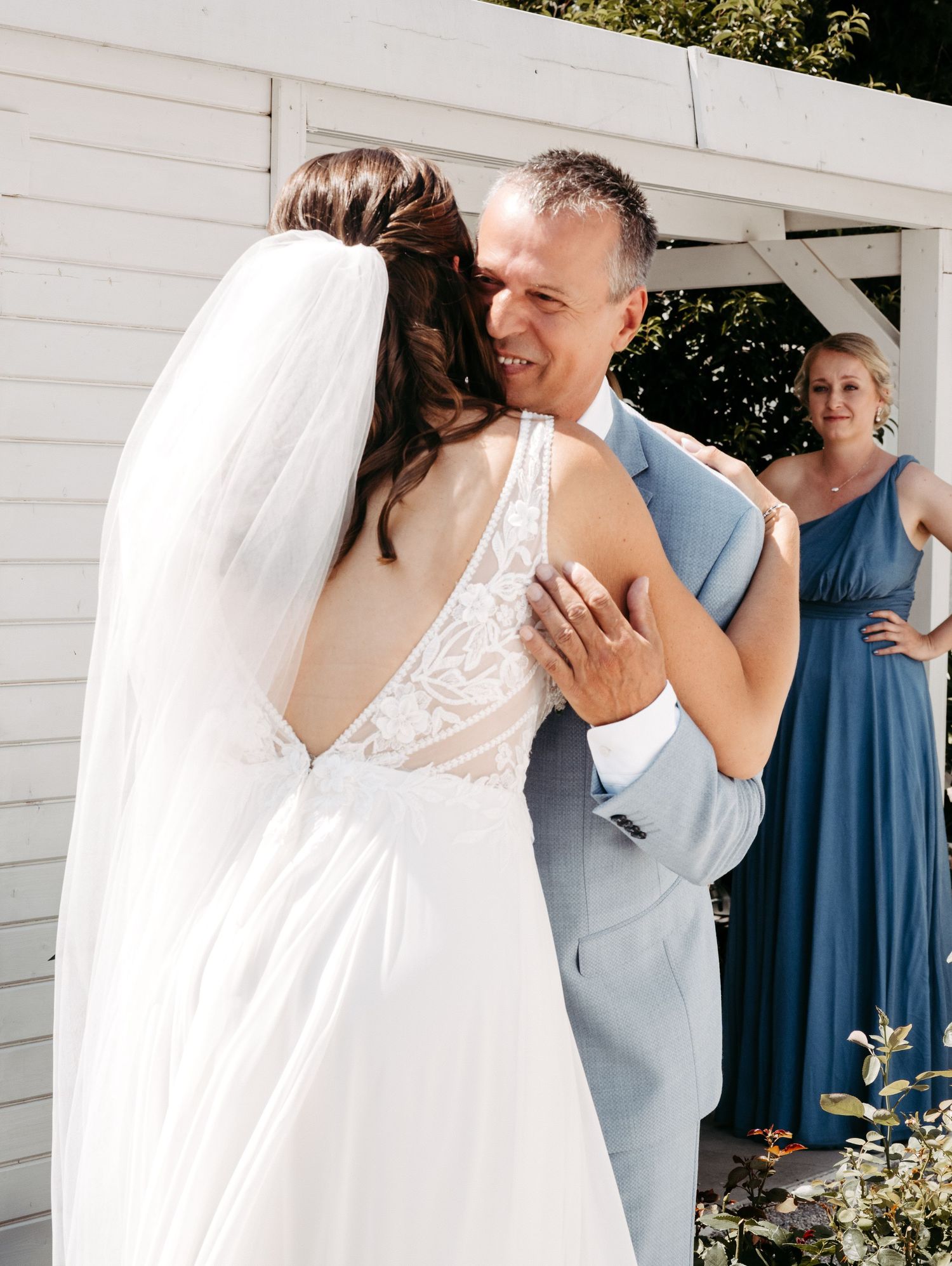 Bride and groom share a warm embrace during outdoor wedding ceremony, bride in lace back gown with veil, groom in light blue suit.