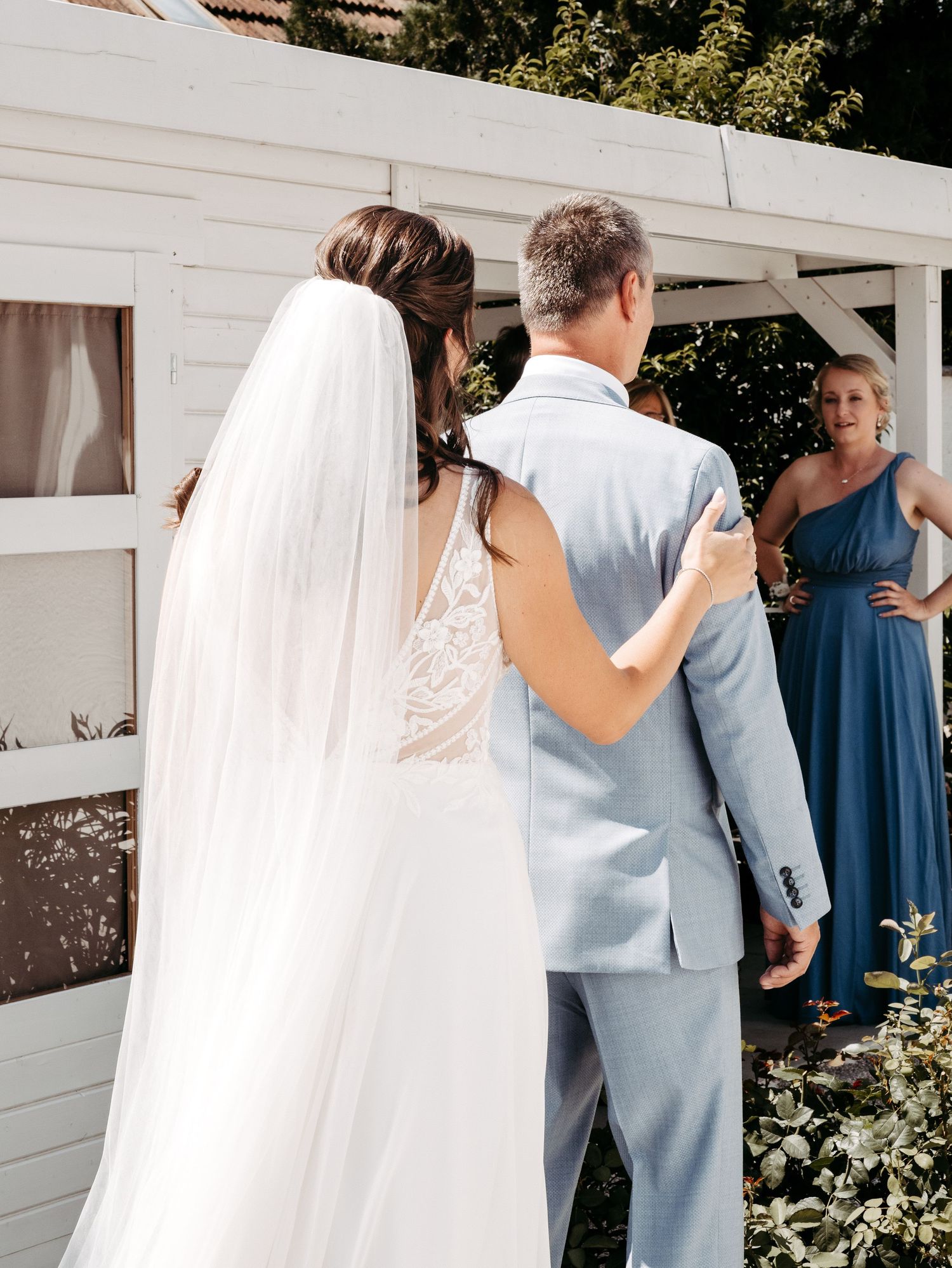 Bride in white gown and veil walks arm-in-arm with man in light blue suit at outdoor wedding ceremony.
