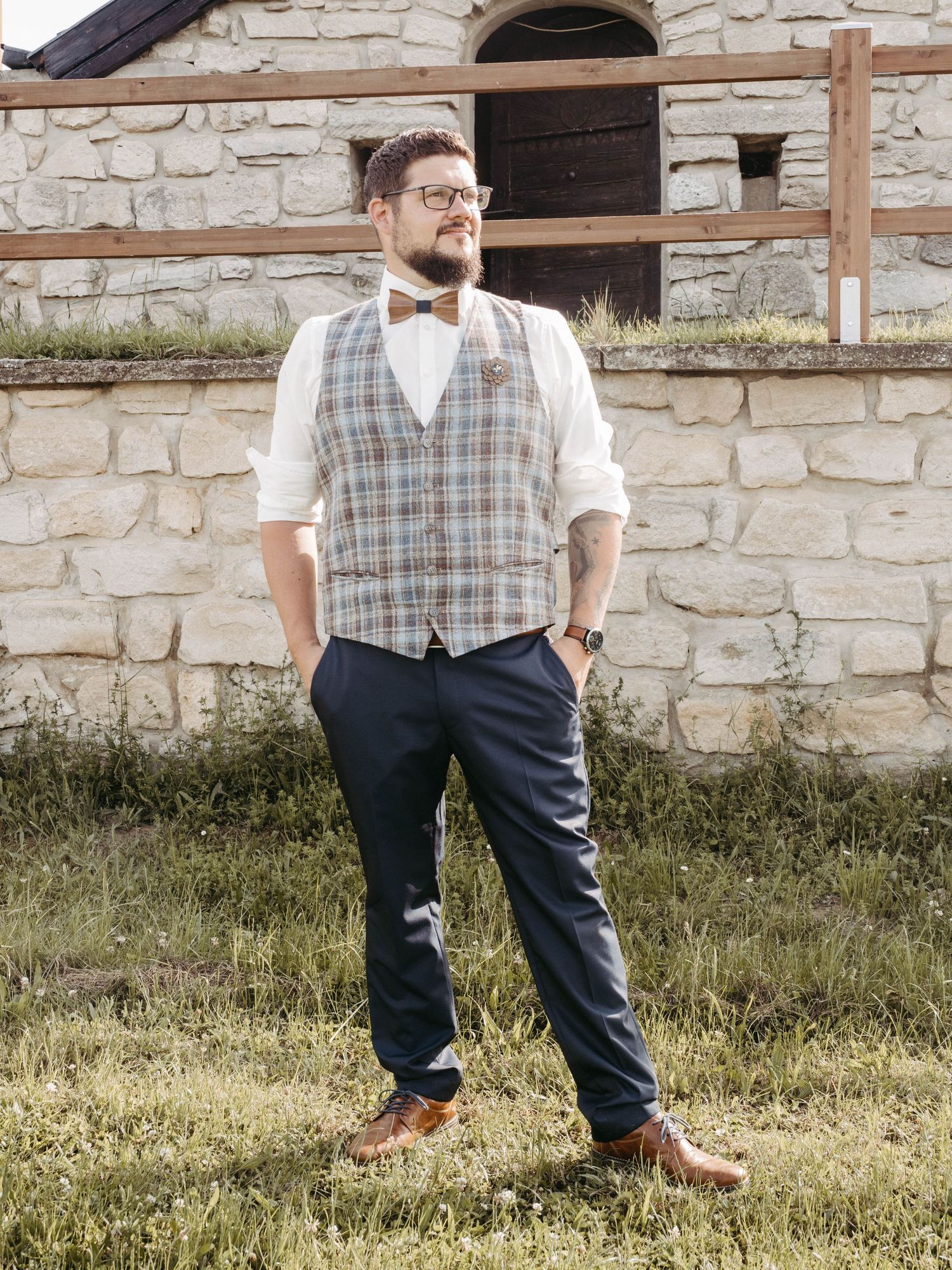 Man in plaid vest, bow tie, white shirt, and navy trousers poses outdoors near a stone wall.
