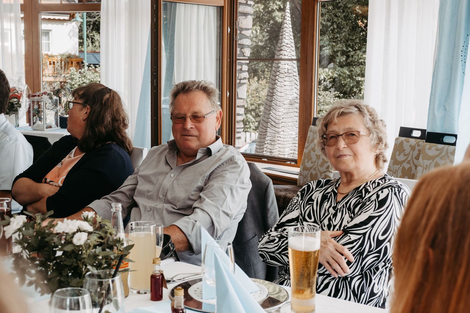 Elderly couple sitting at a festive dining table during a celebration, with drinks and floral decorations nearby.