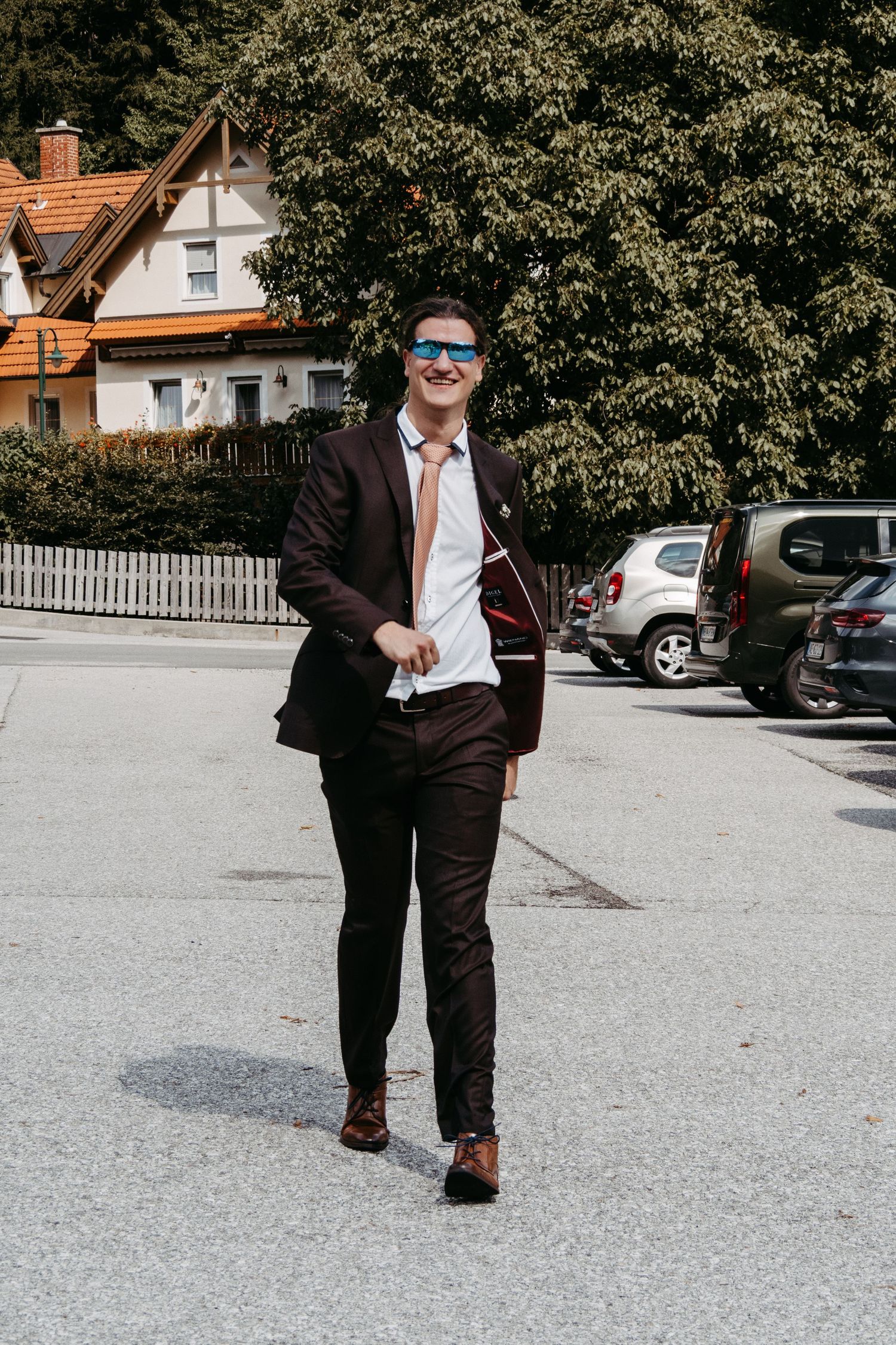 Smiling man in dark burgundy suit and blue sunglasses walking outdoors near parked cars and houses.