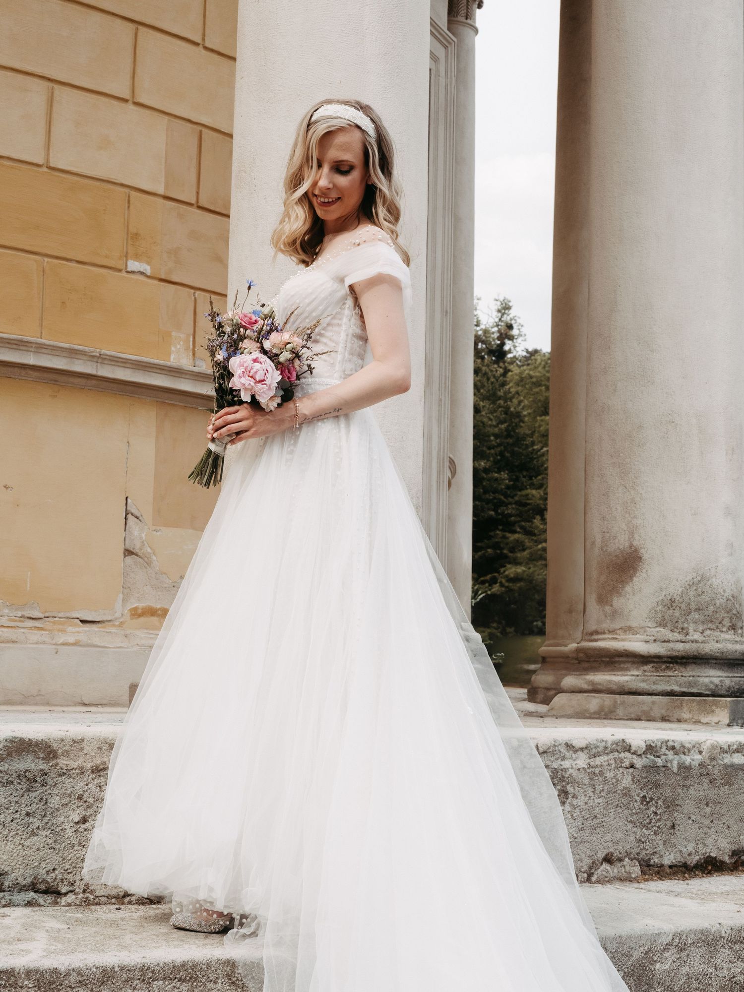 Bride in flowing white tulle gown posing gracefully near classical stone columns.