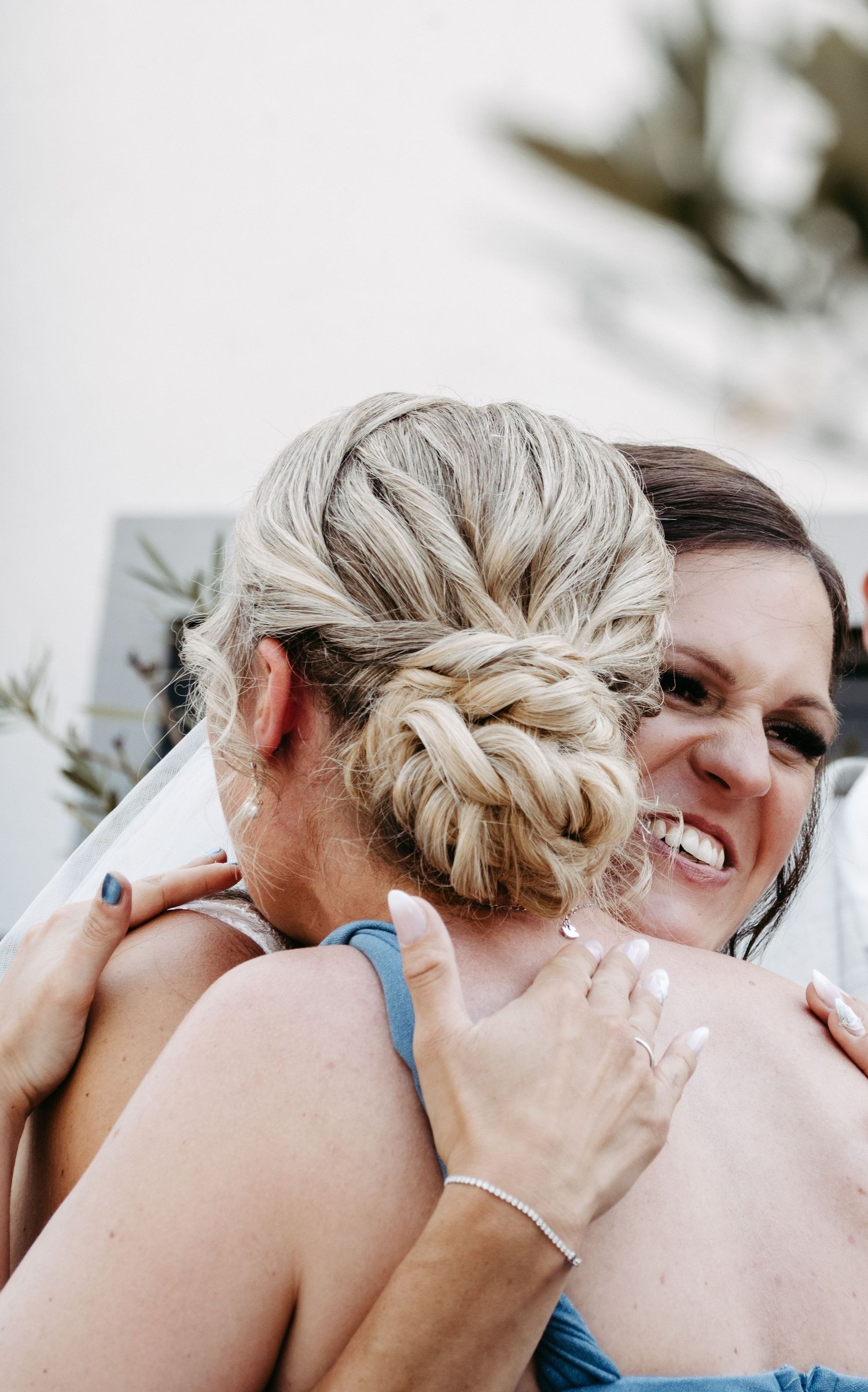 Two women sharing a warm hug, one with an elegant braided updo hairstyle, wearing blue dresses outdoors.