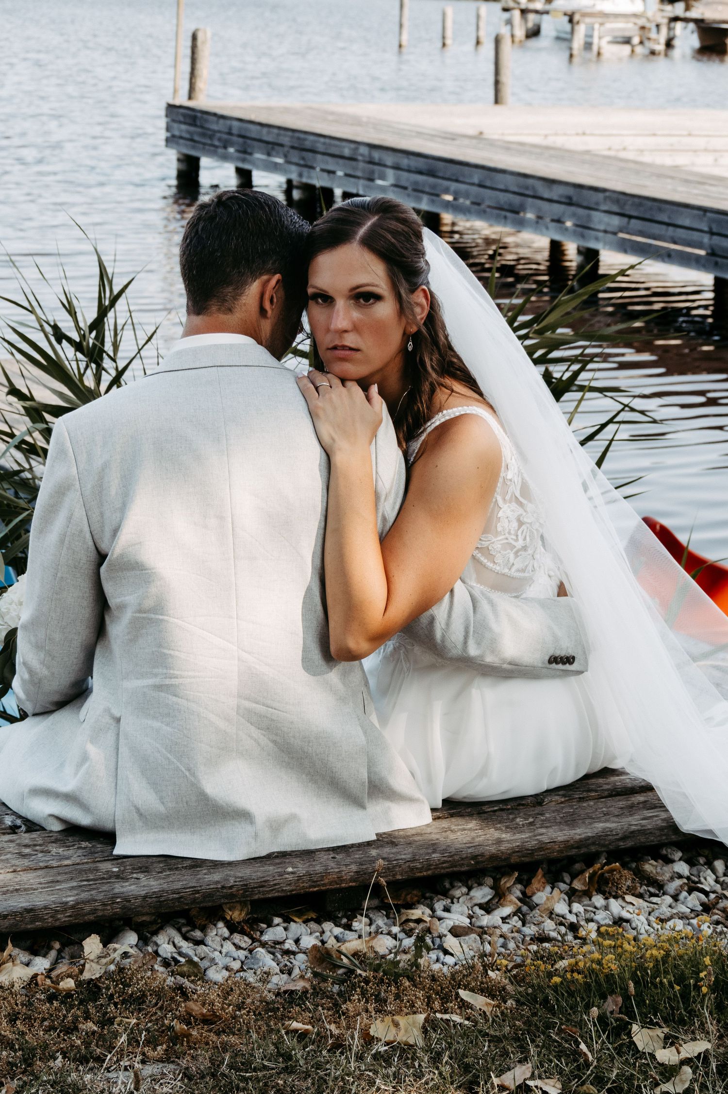 Bride in white gown and veil sits with groom by waterfront dock during romantic outdoor wedding photo session.