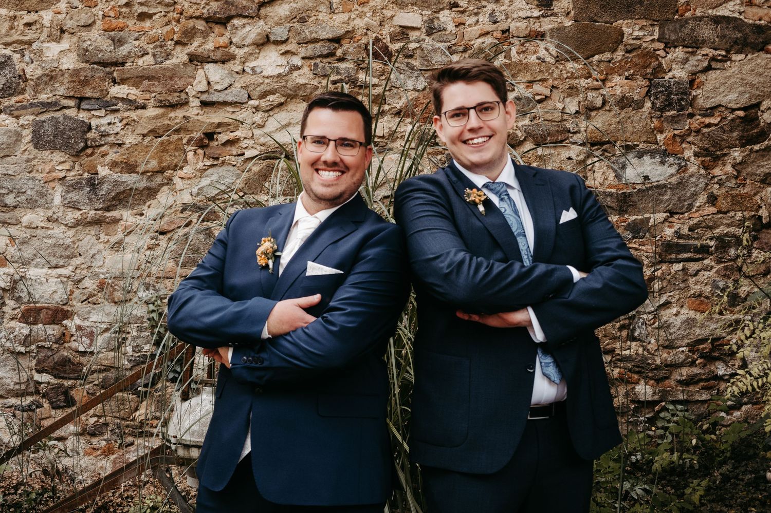 Two groomsmen in navy suits posing back-to-back against rustic stone wall.