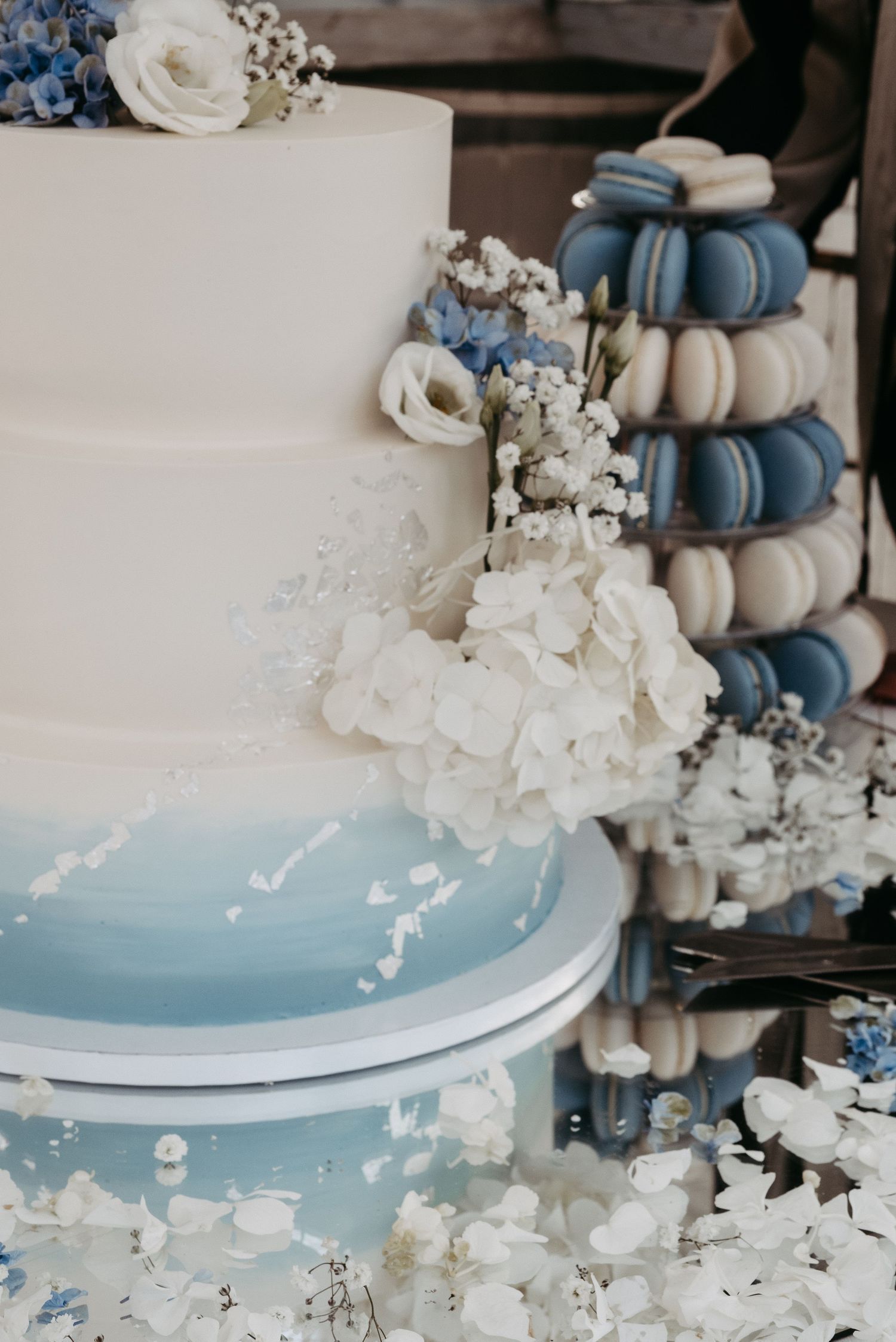 Elegant three-tier white and blue ombre wedding cake adorned with white hydrangeas beside a tower of blue macarons.