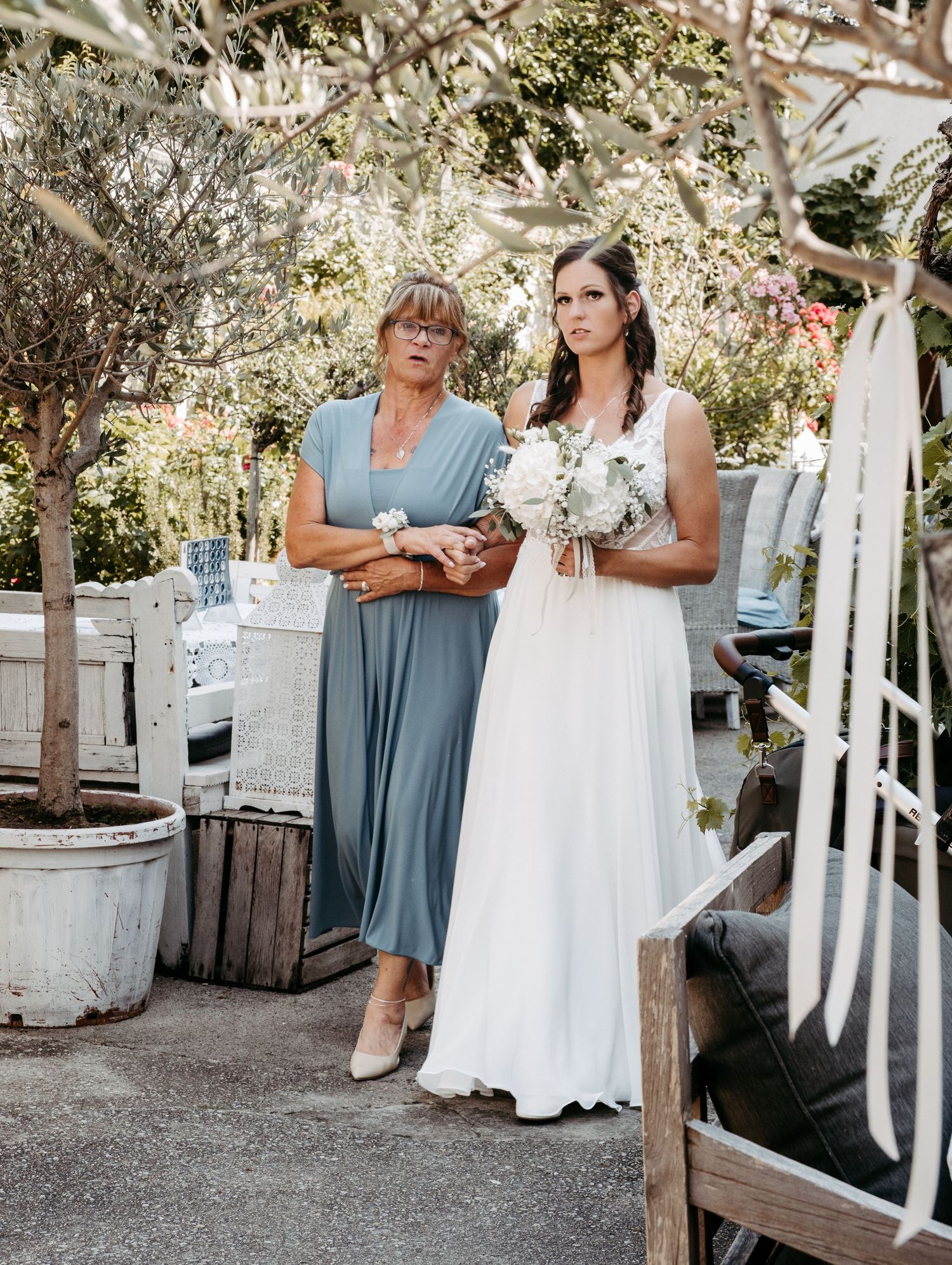 Bride in white gown holding bouquet stands with older woman in blue dress at outdoor wedding venue.