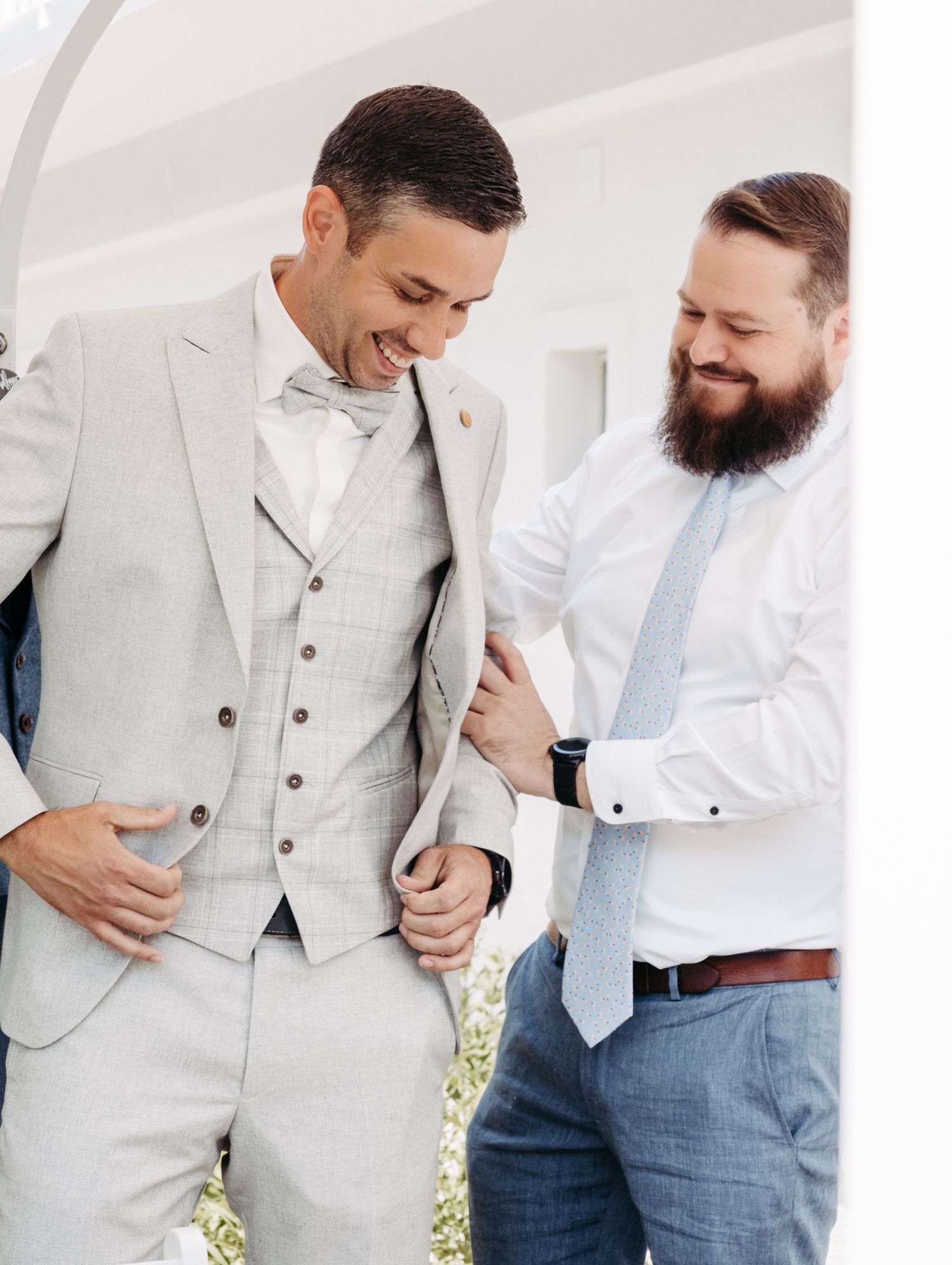 Groom in light gray three-piece suit with bow tie being helped by bearded groomsman in white shirt at wedding.