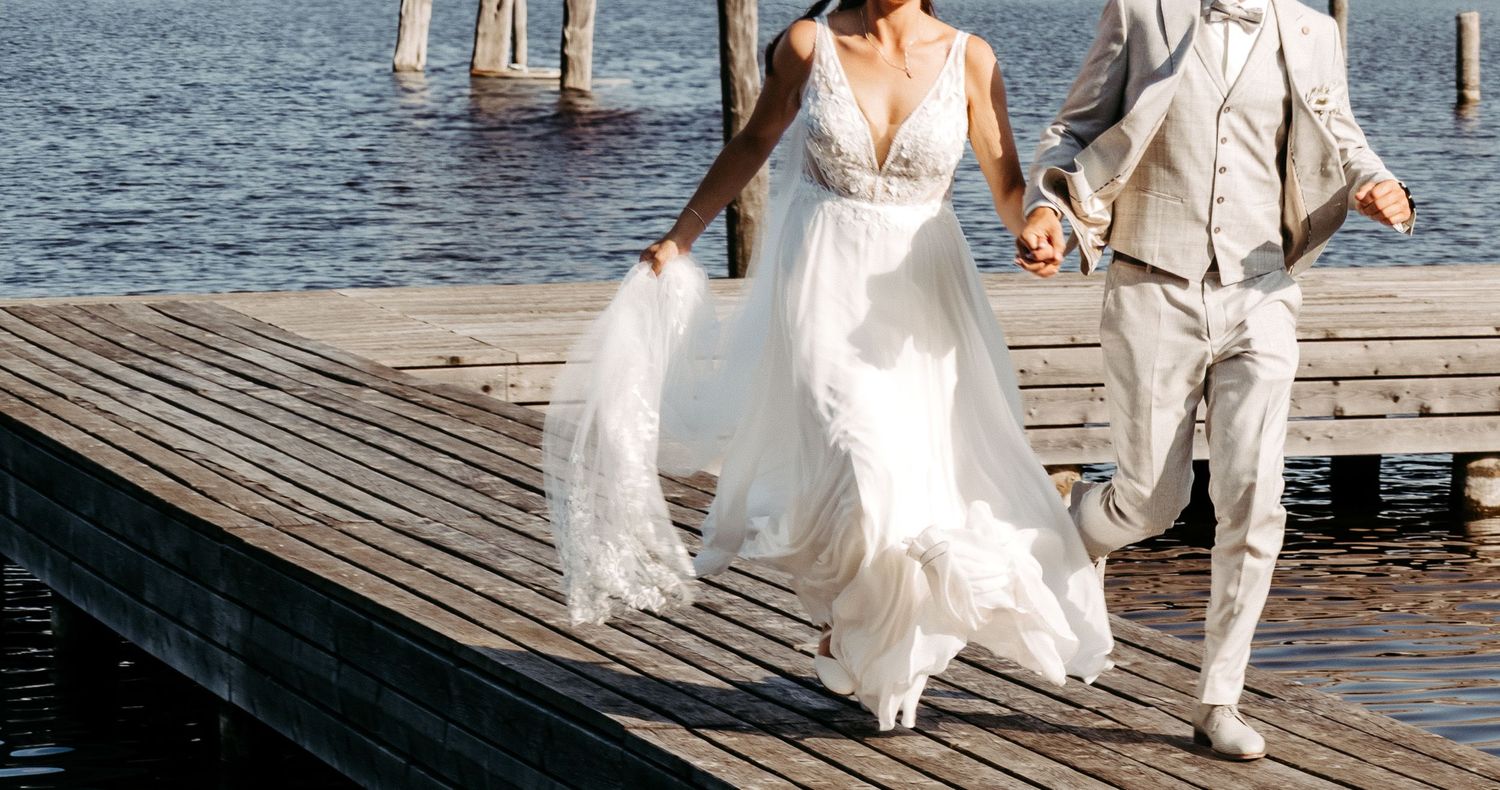Bride in white gown and groom in beige suit running hand-in-hand on a wooden dock by the water.