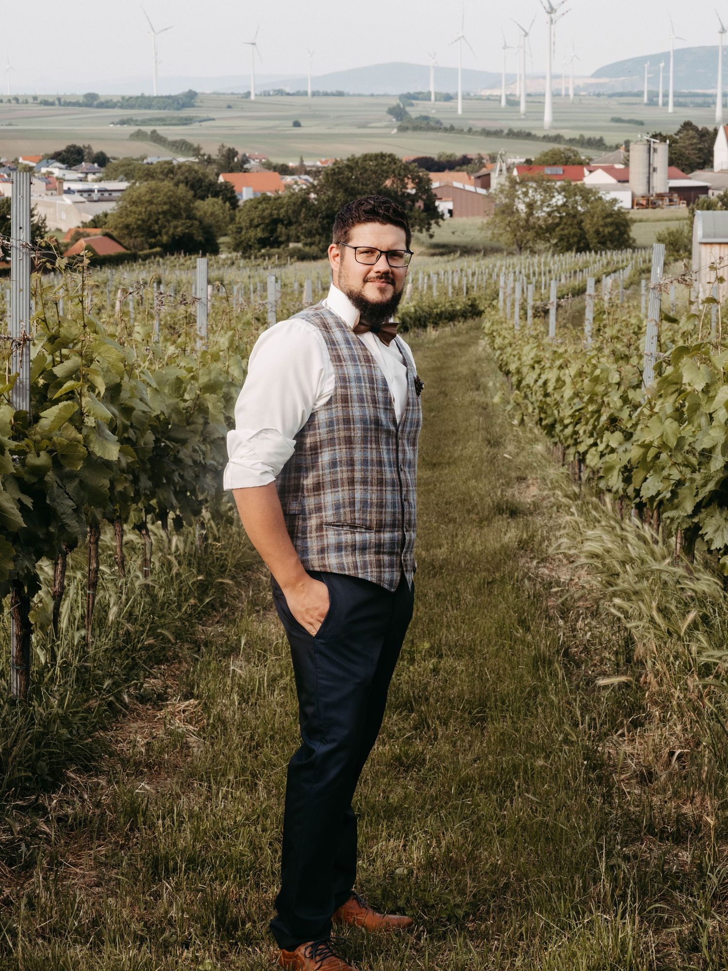 Man in plaid vest stands among vineyard rows with wind turbines and village in background.