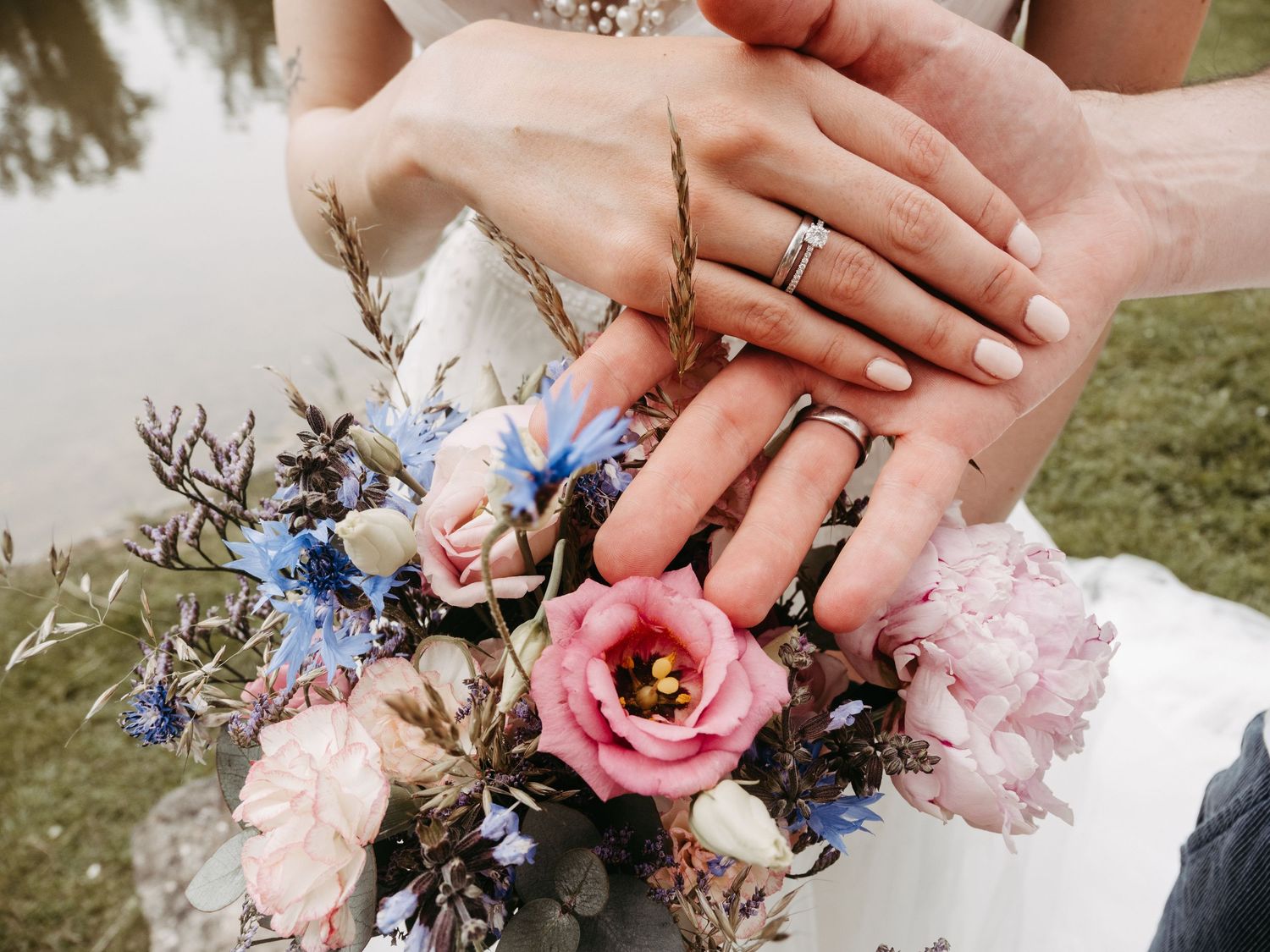 Newlywed couple displaying wedding rings over a colorful bridal bouquet with pink and blue flowers.