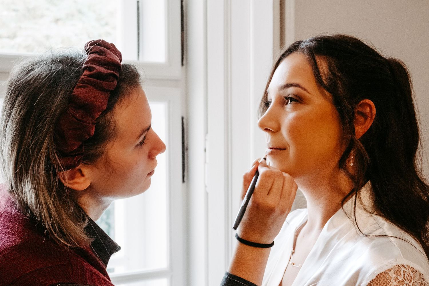 A makeup artist applying makeup to a bride's face near a bright window during wedding preparations.