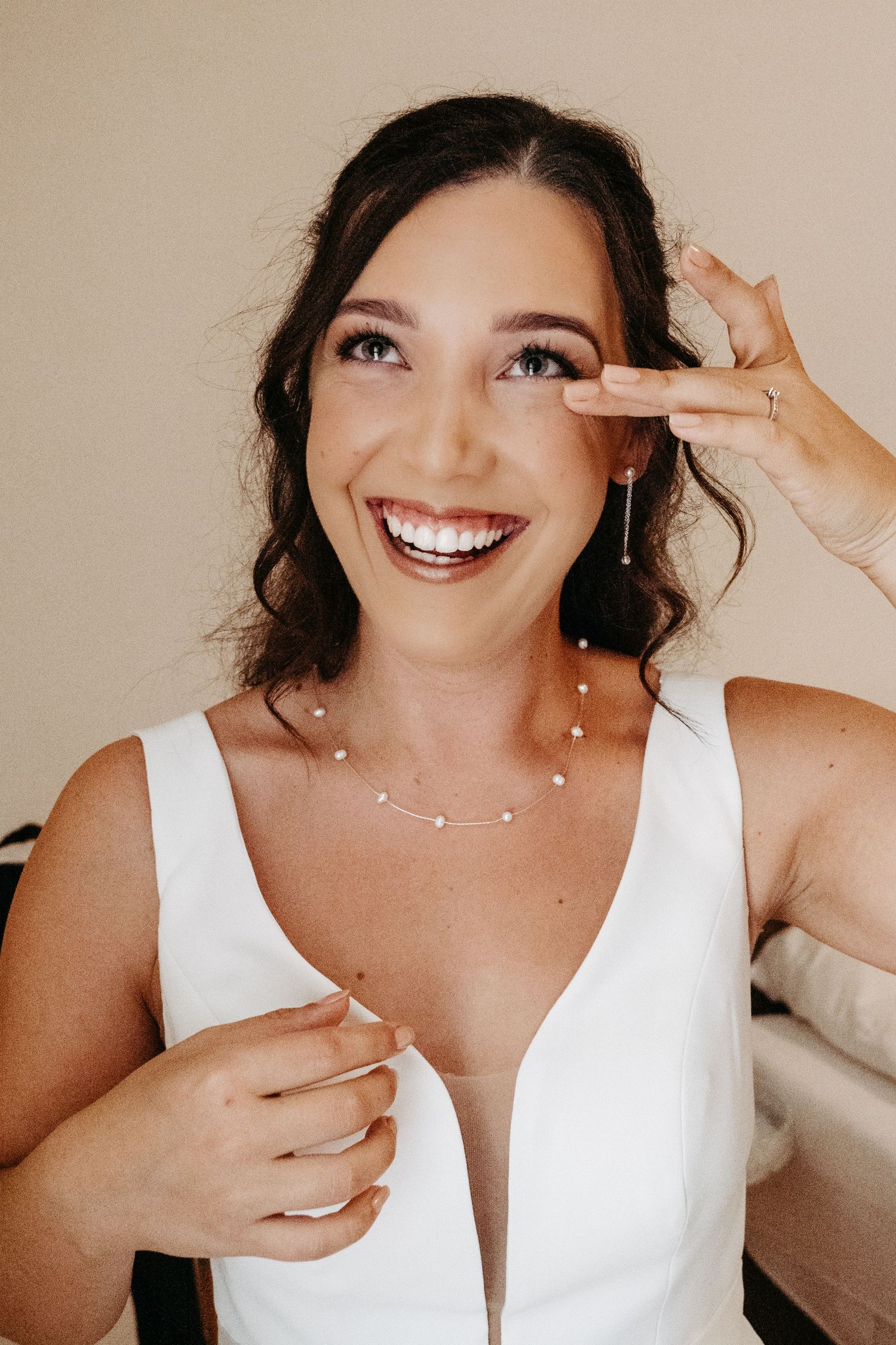 Smiling bride in white dress wearing pearl necklace and earrings touching her face during wedding preparation.