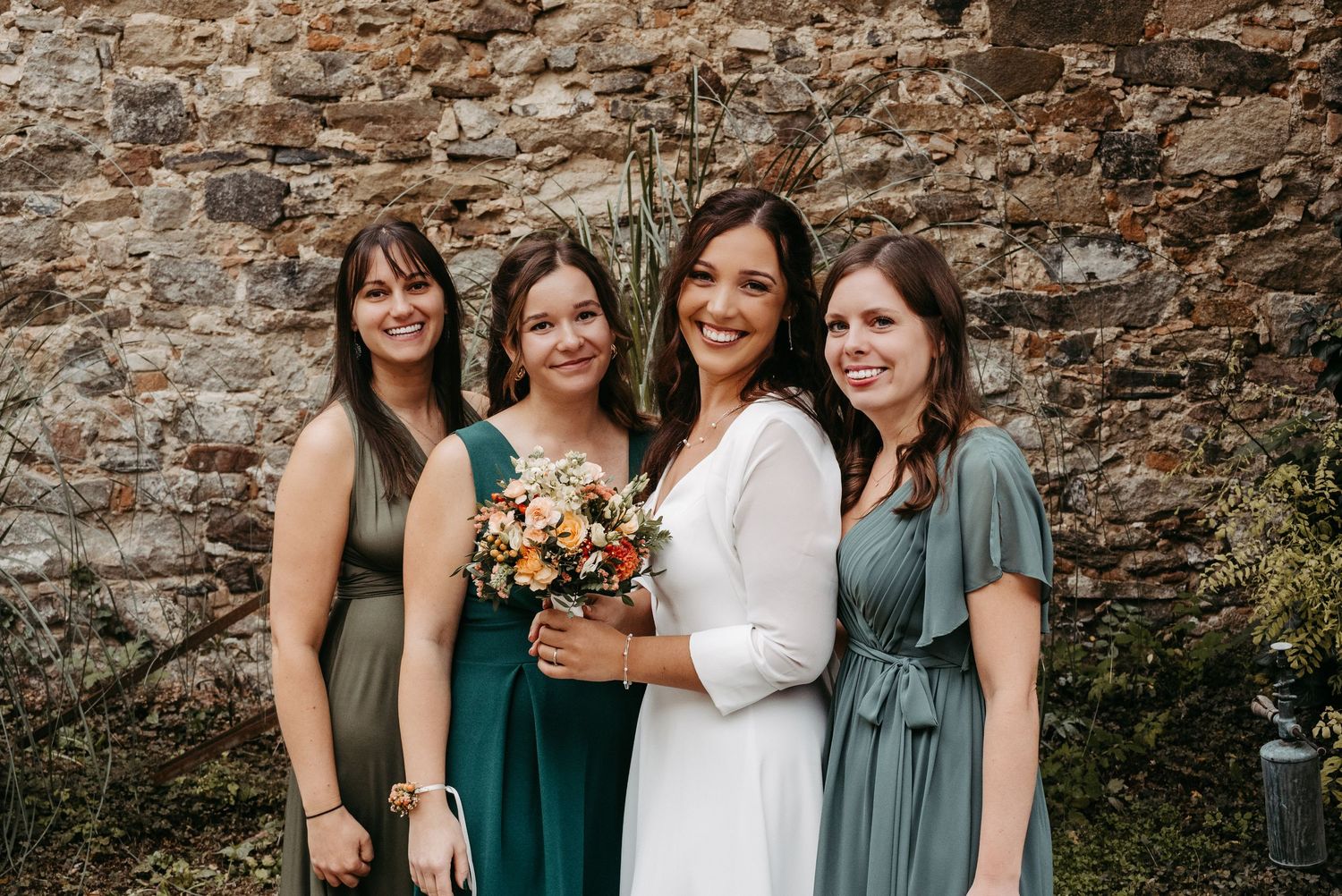 Bride in white dress poses with three bridesmaids in green dresses holding floral bouquet outdoors.