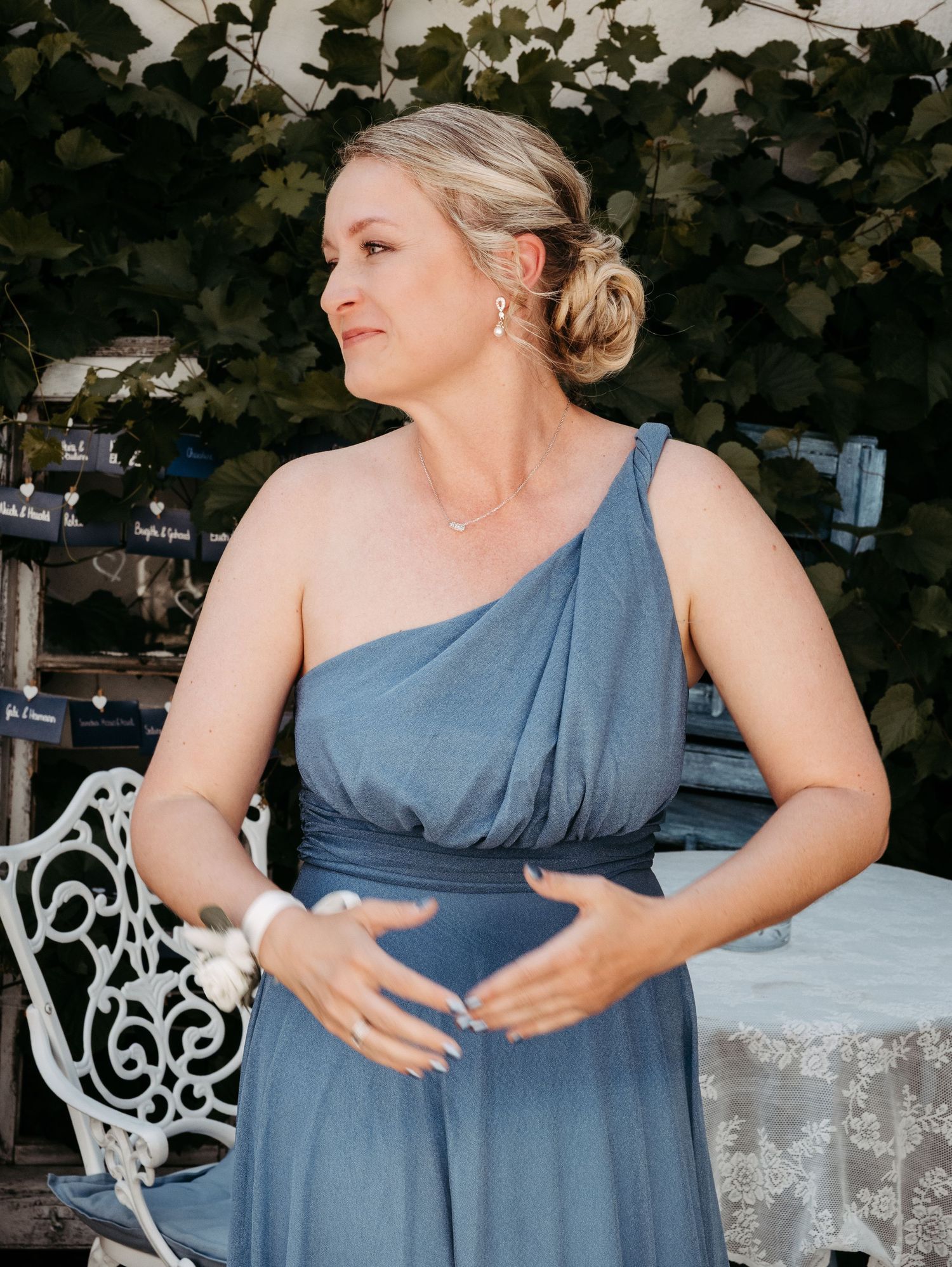 Pregnant woman in blue one-shoulder dress cradling baby bump at outdoor wedding venue.
