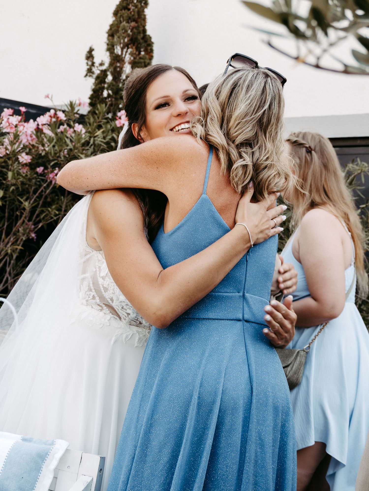 A bride in a white lace gown and veil shares a warm hug with a guest in a blue dress at an outdoor wedding.