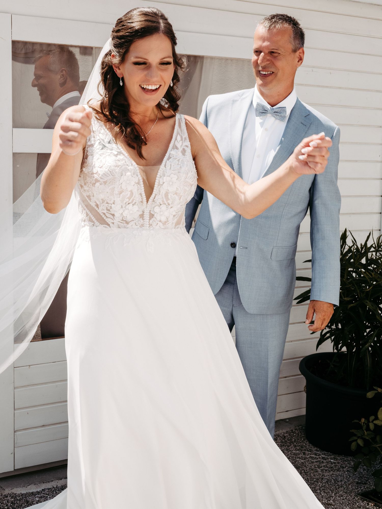 Bride in lace V-neck wedding gown twirls veil as groom in light blue suit smiles behind her outdoors.