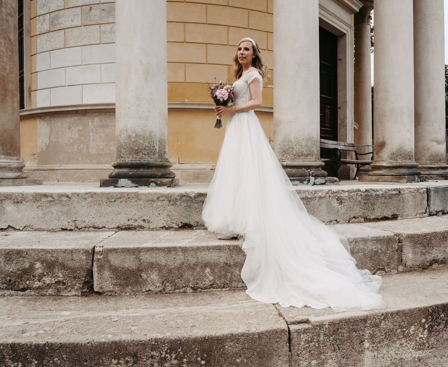 Bride in elegant white gown with train poses on stone steps holding bouquet near classical columns.