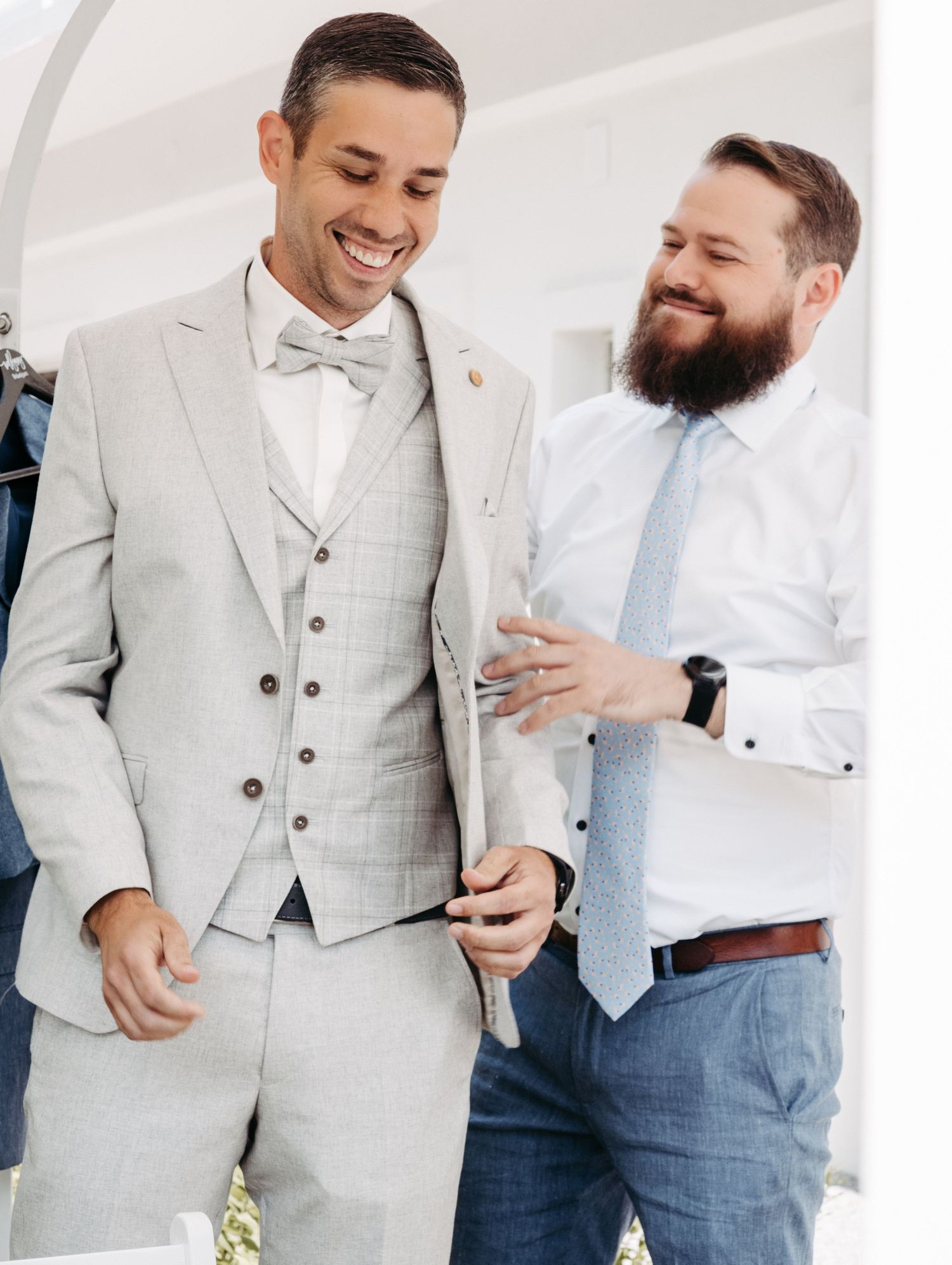 Two men in formal attire smile together, one wearing a light gray three-piece suit with bow tie at a wedding.