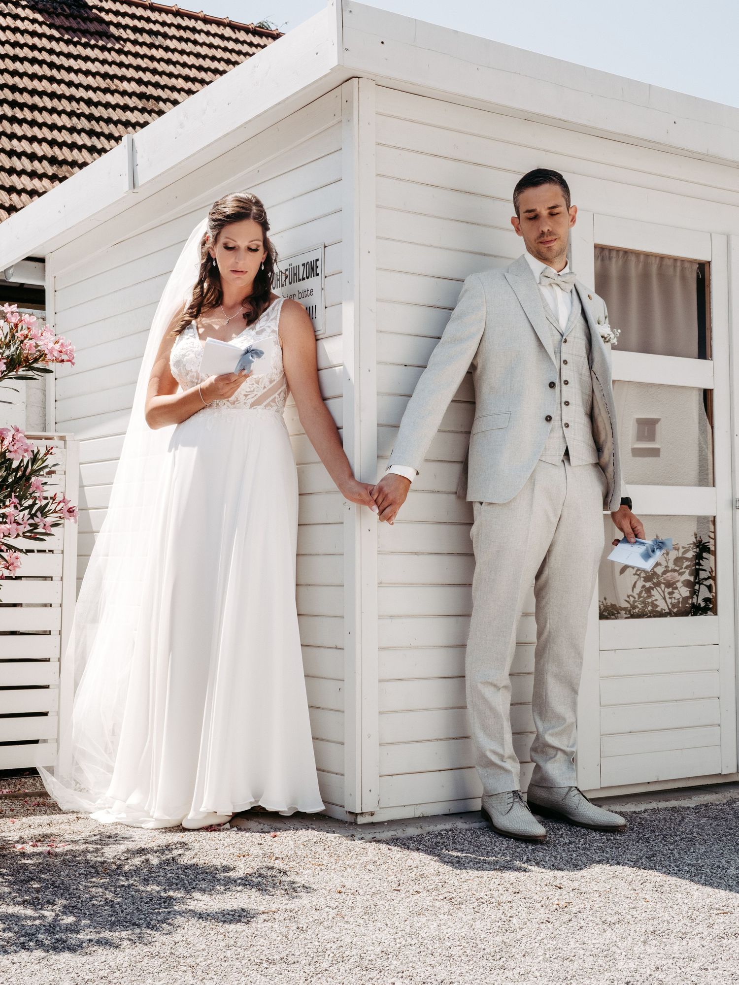 Bride in white gown and groom in beige suit hold hands leaning against a white wooden building on their wedding day.