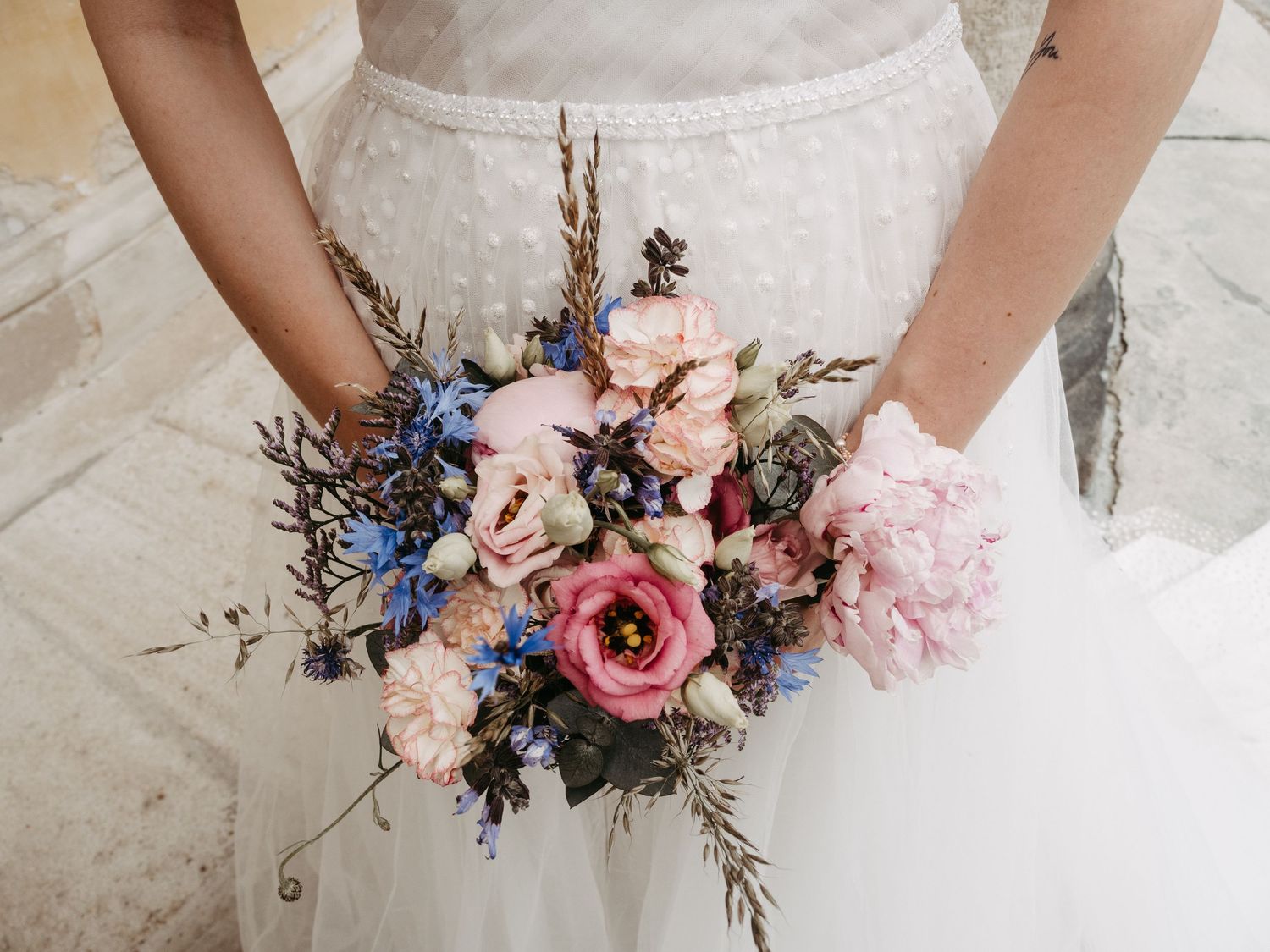 Wedding bouquet featuring pink roses and blue flowers held against white bridal gown.
