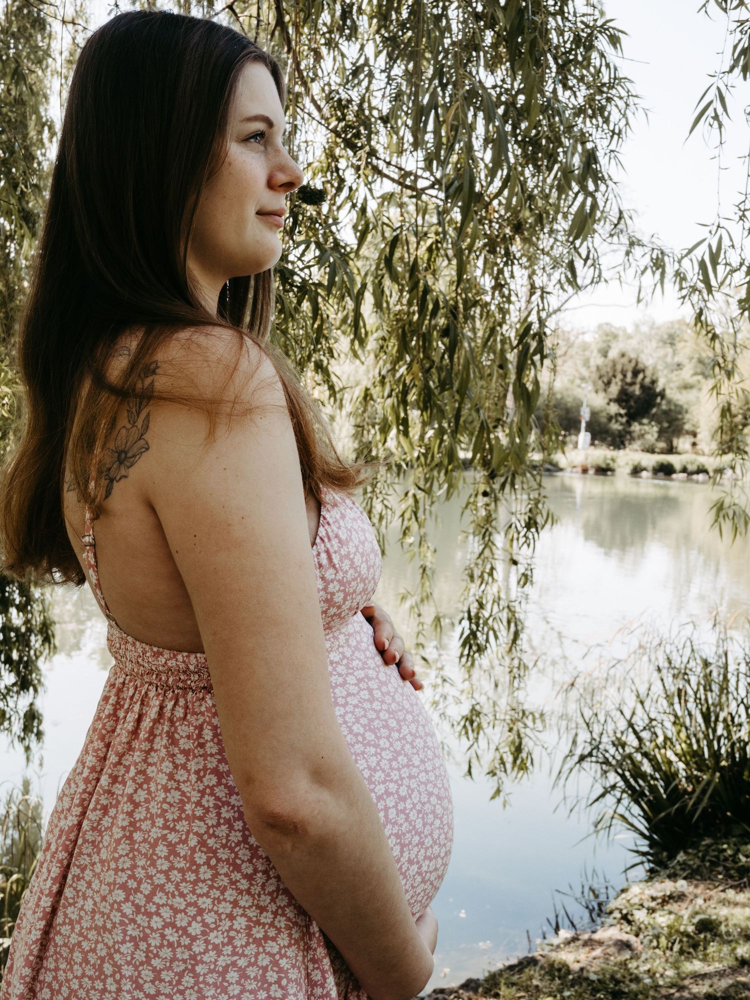 Maternity photoshoot by a lake with willow trees in soft natural lighting.