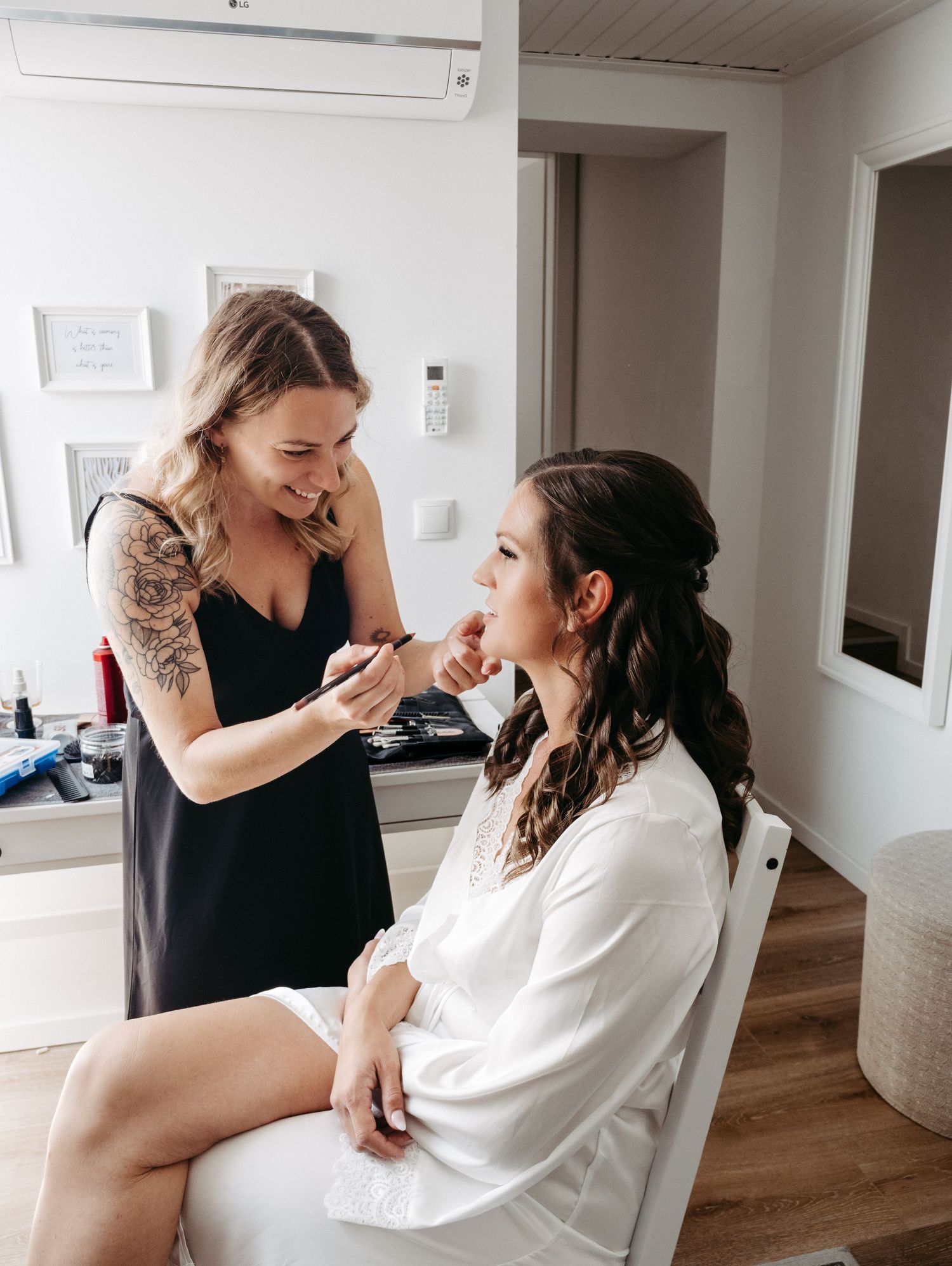 A makeup artist with tattoos applies makeup to a bride in a white robe during wedding preparations.