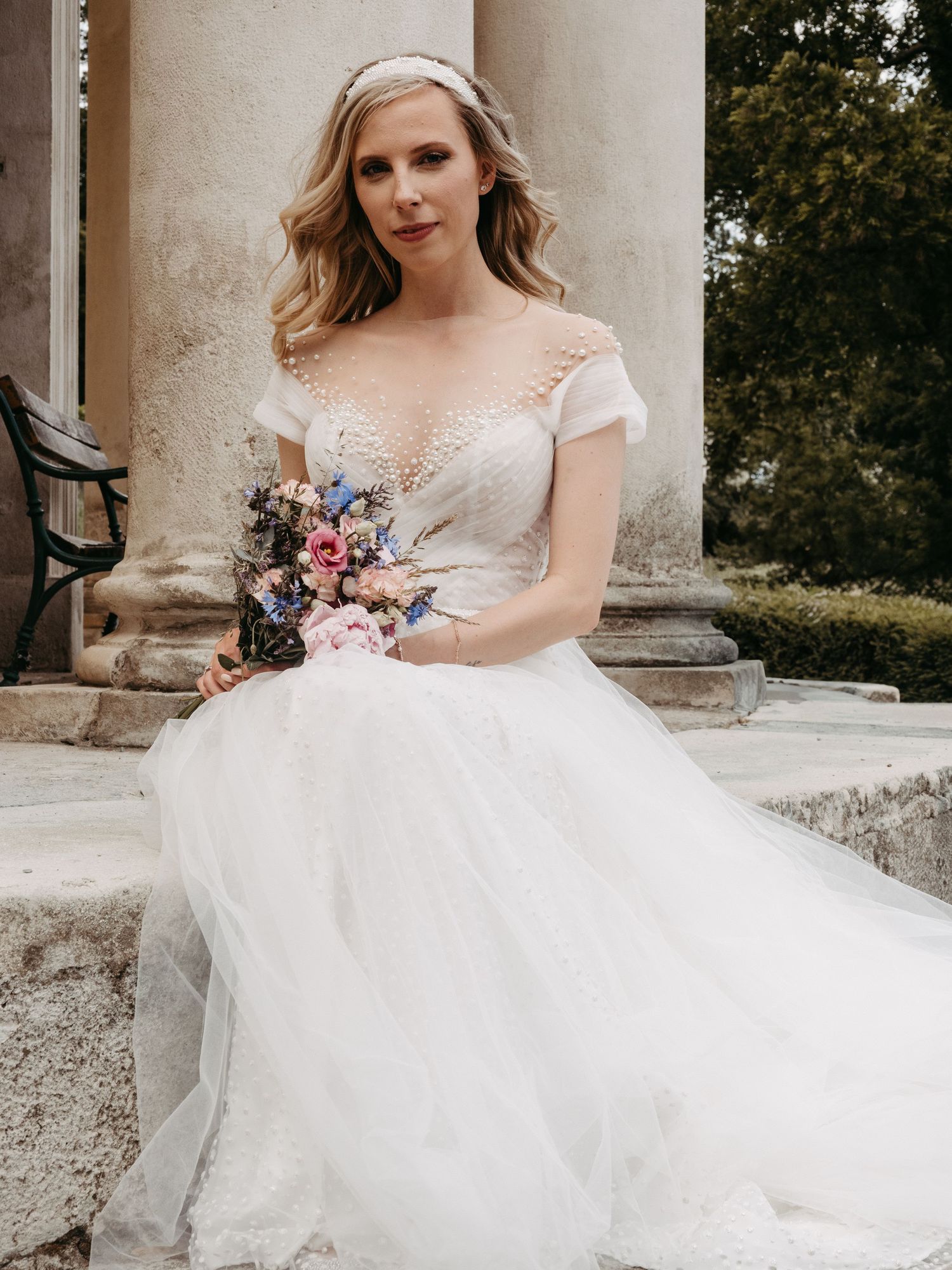 Bride in white tulle ballgown with floral bodice holds colorful bouquet, seated by stone columns outdoors.