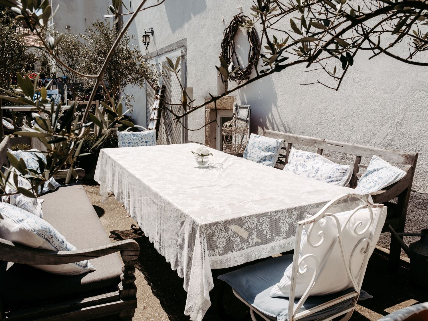 Charming outdoor patio with white lace tablecloth, wooden chairs, and Mediterranean-style decor in bright sunlight.