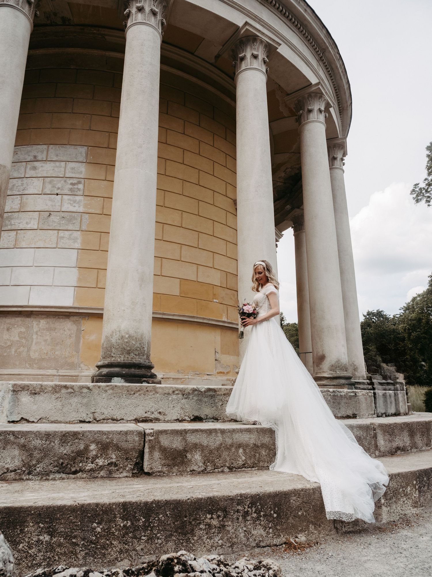 Bride in white gown holding bouquet stands on stone steps of classical columned building on cloudy day.
