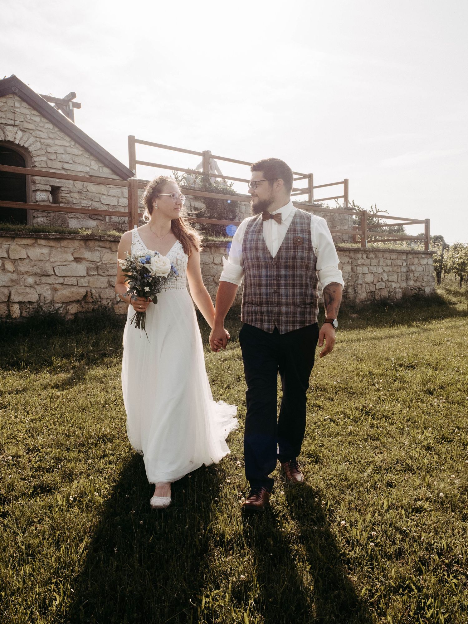 Bride and groom holding hands walking on green grass near a stone building on their wedding day.