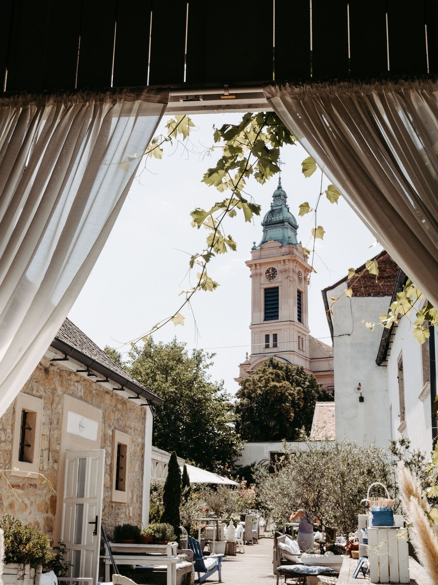 Charming European courtyard with white curtains framing a view of a historic church tower surrounded by lush greenery.