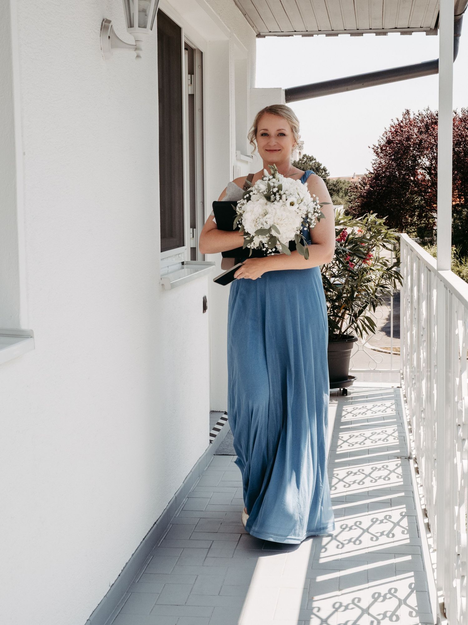 Bridesmaid in dusty blue gown holding white floral bouquet on sunlit balcony.
