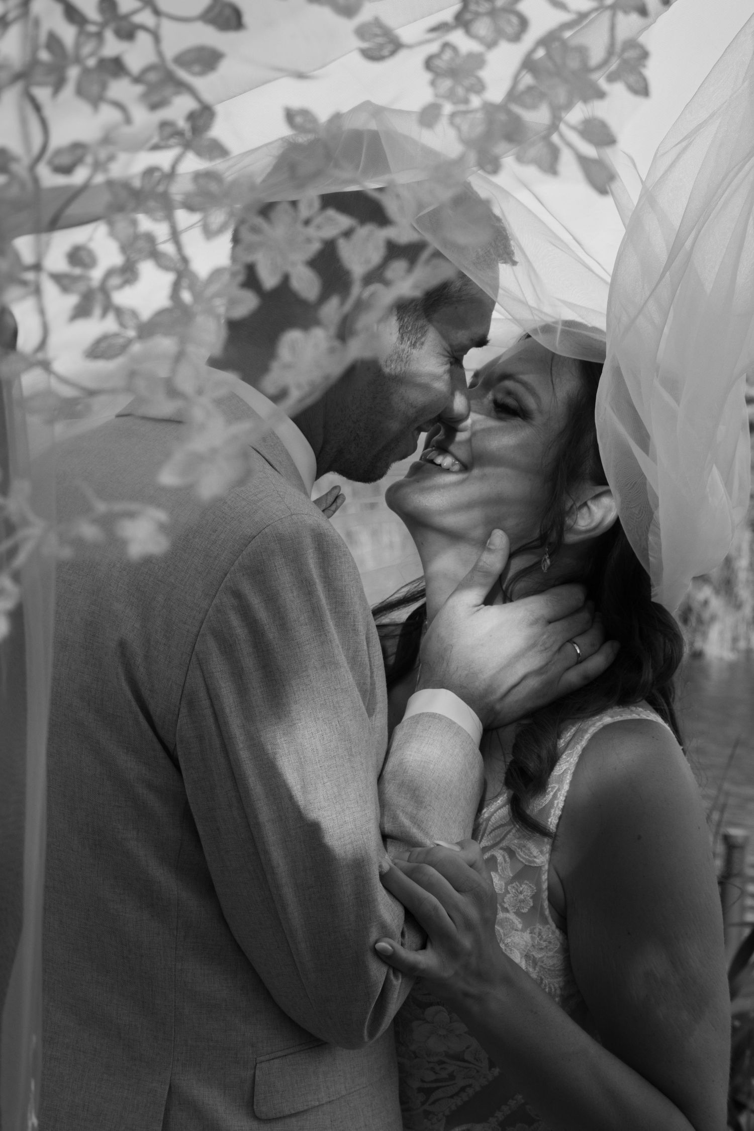 Black and white photo of a bride and groom sharing a romantic kiss under a floral wedding veil.