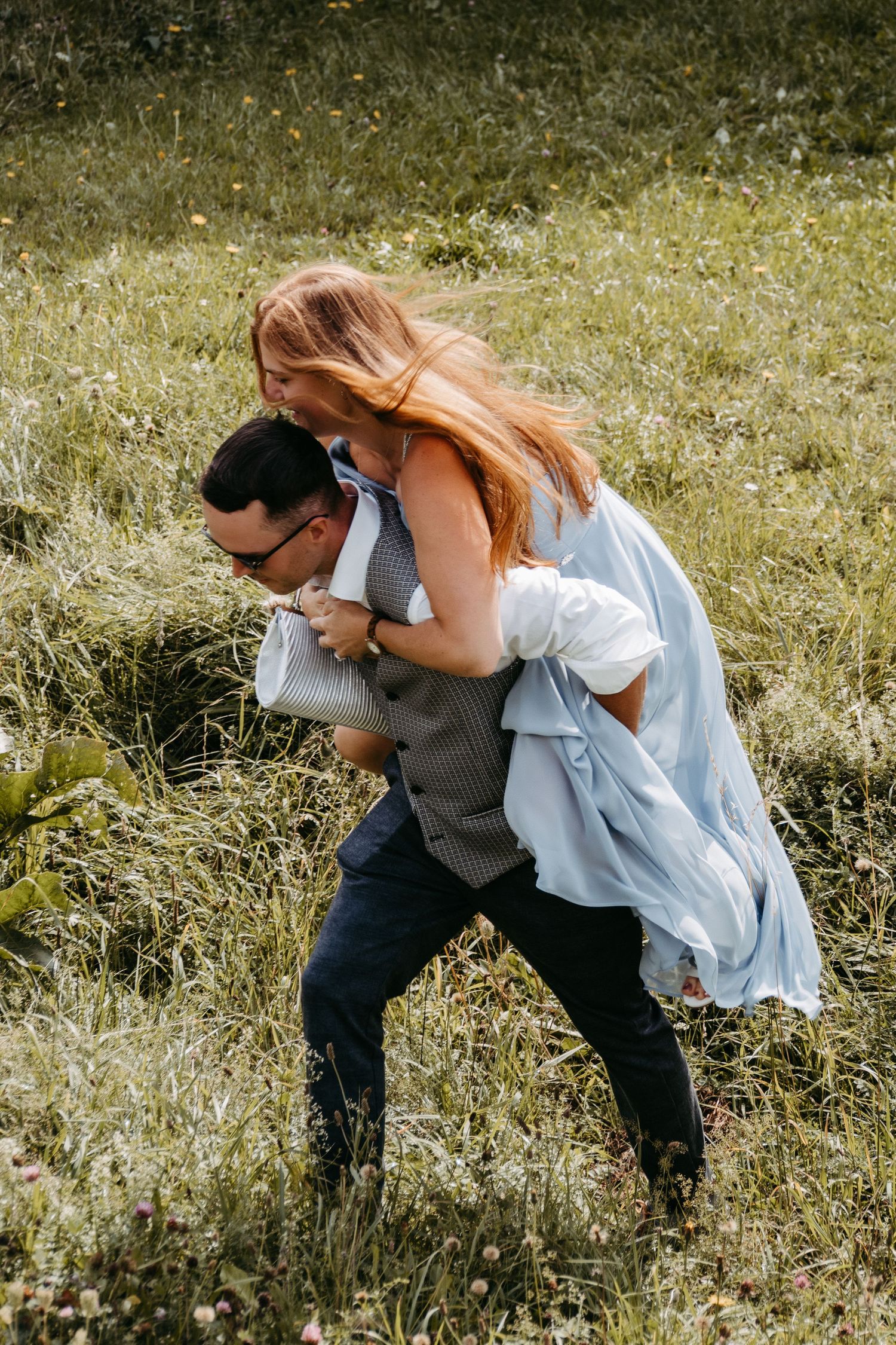 Man giving woman a piggyback ride in a grassy meadow, both laughing and dressed in elegant casual attire.