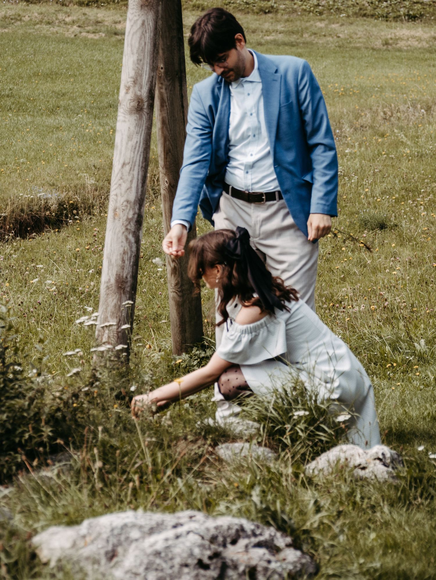 A man in a blue blazer assists a woman in a light dress picking flowers in a green meadow near a wooden post.