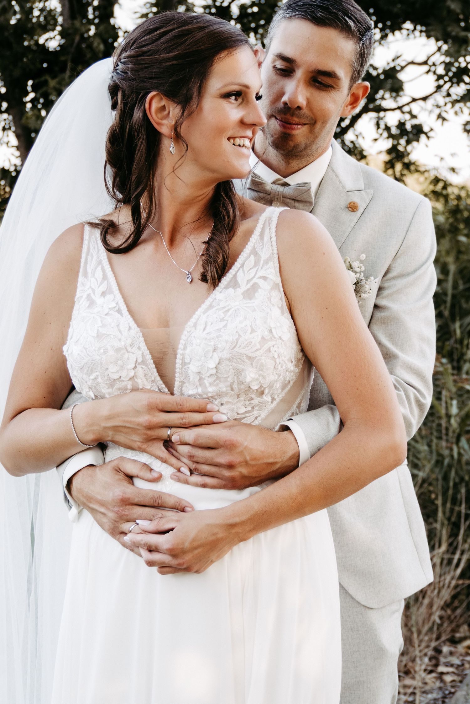 Bride in white lace gown and veil embraced by groom in beige suit, sharing a romantic outdoor wedding moment.