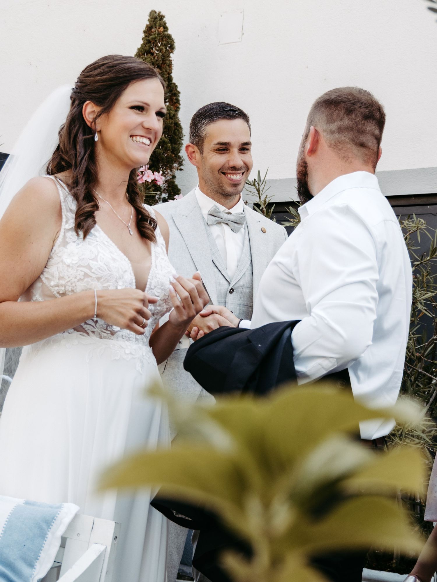 Bride and groom exchanging rings during outdoor wedding ceremony with smiling officiant in light gray suit.