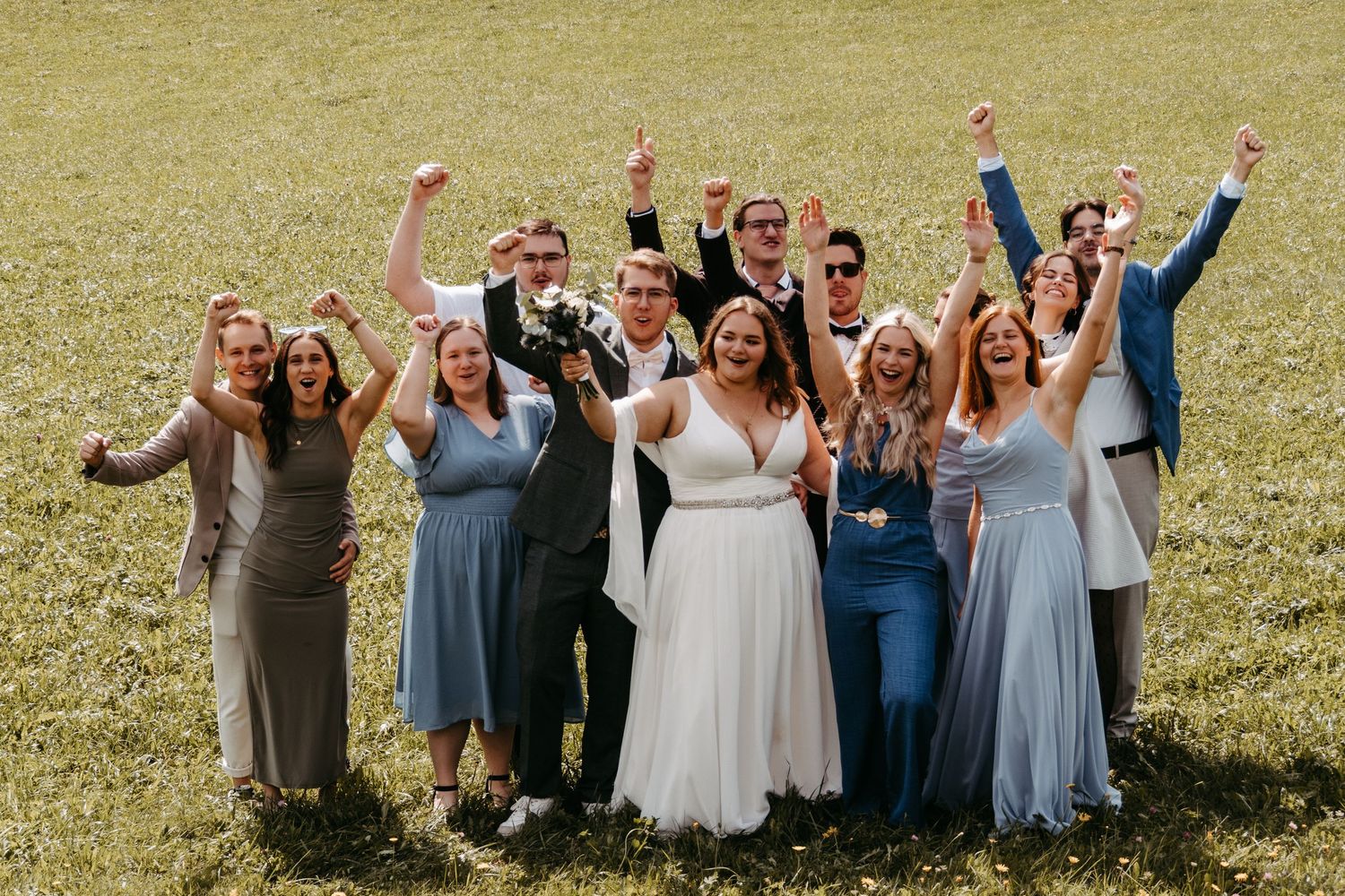 Wedding party celebrating outdoors on green grass, bride in white gown surrounded by cheering guests in blue and formal attire.