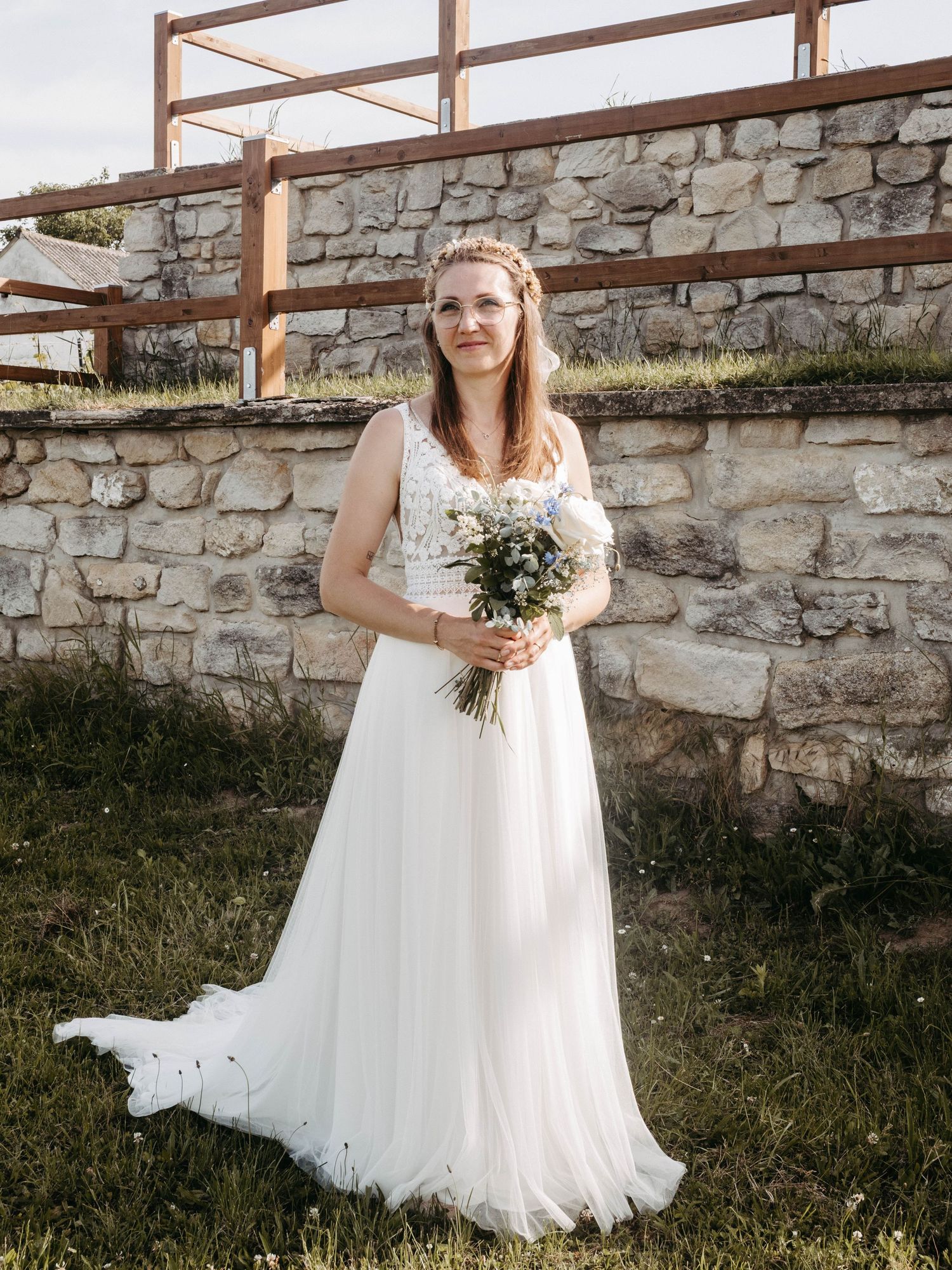 Bride in white lace wedding gown holding wildflower bouquet, standing before rustic stone wall outdoors.