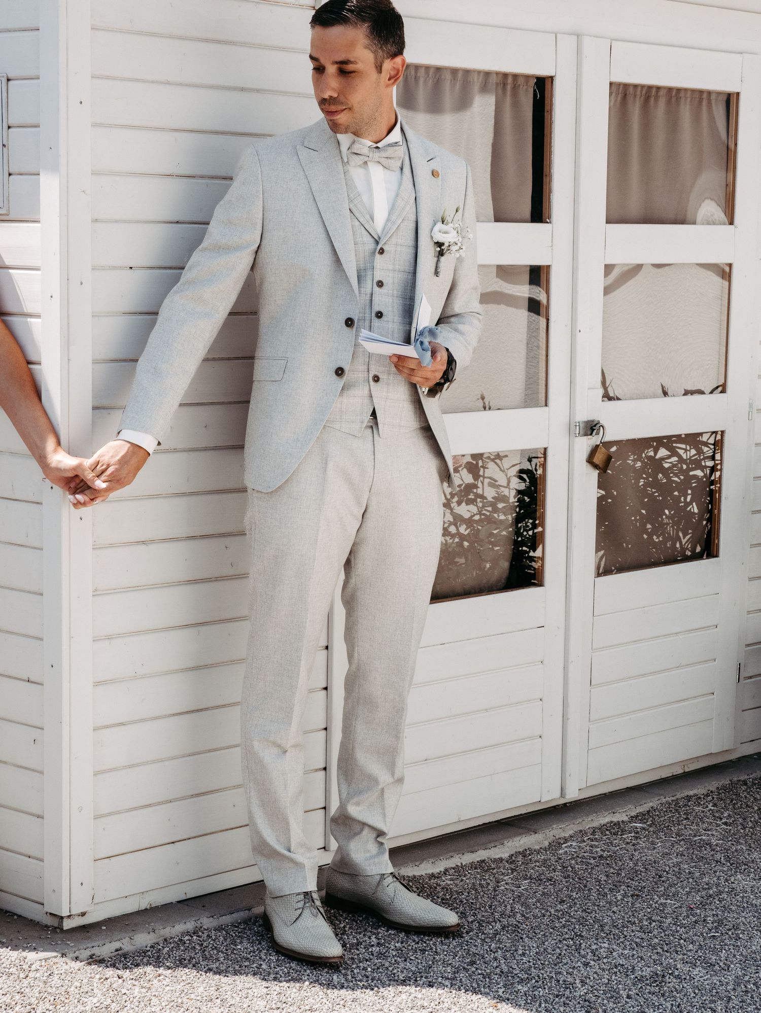 Groom in light gray suit with bow tie and boutonniere stands confidently outside white building on wedding day.