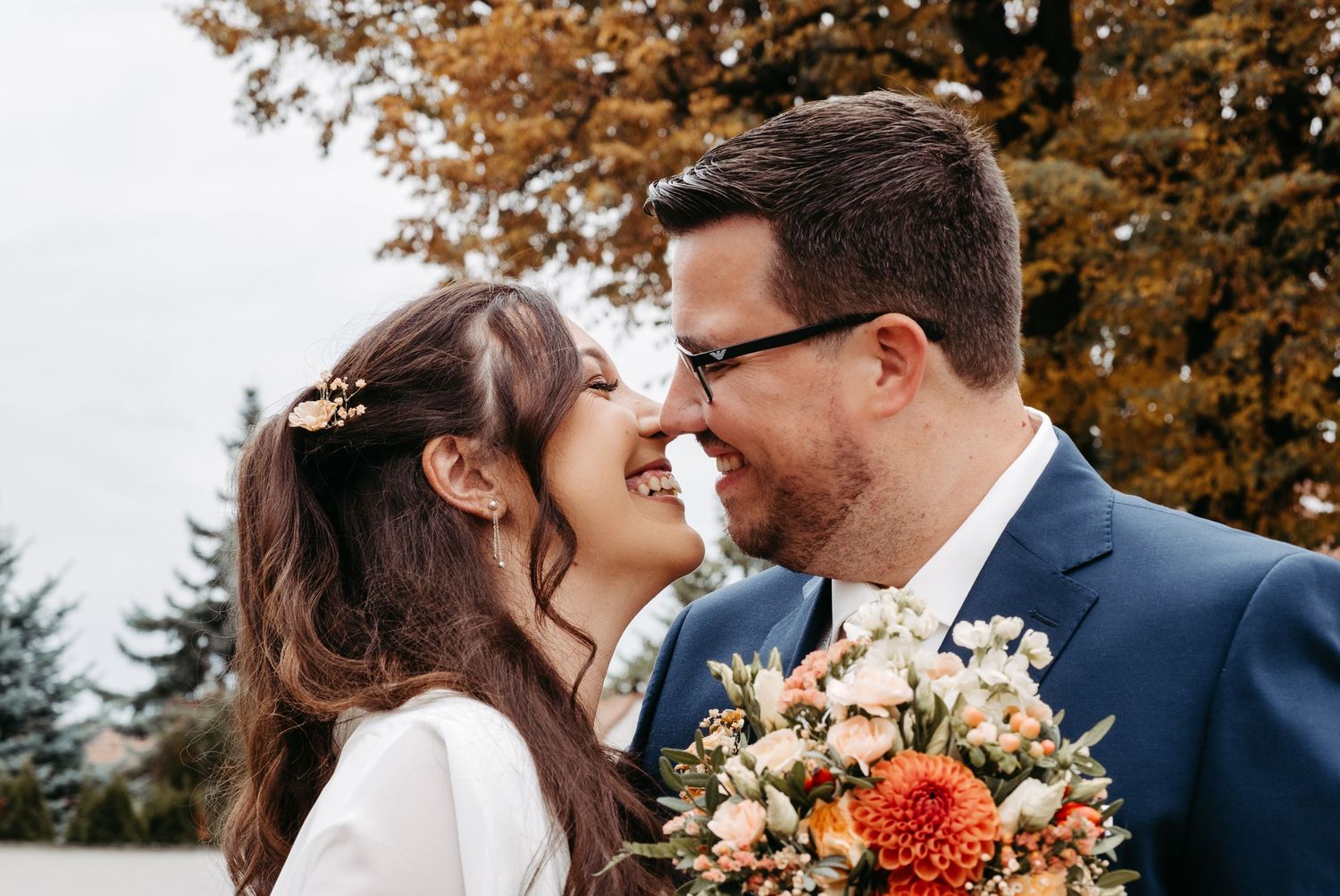Bride and groom sharing a romantic nose-to-nose moment outdoors, with autumn foliage and a colorful bouquet.