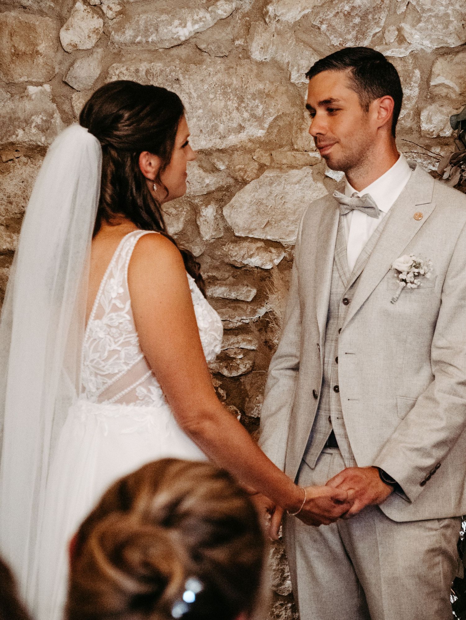 Bride and groom holding hands at wedding altar in front of rustic stone wall, groom in gray suit, bride in white lace gown.