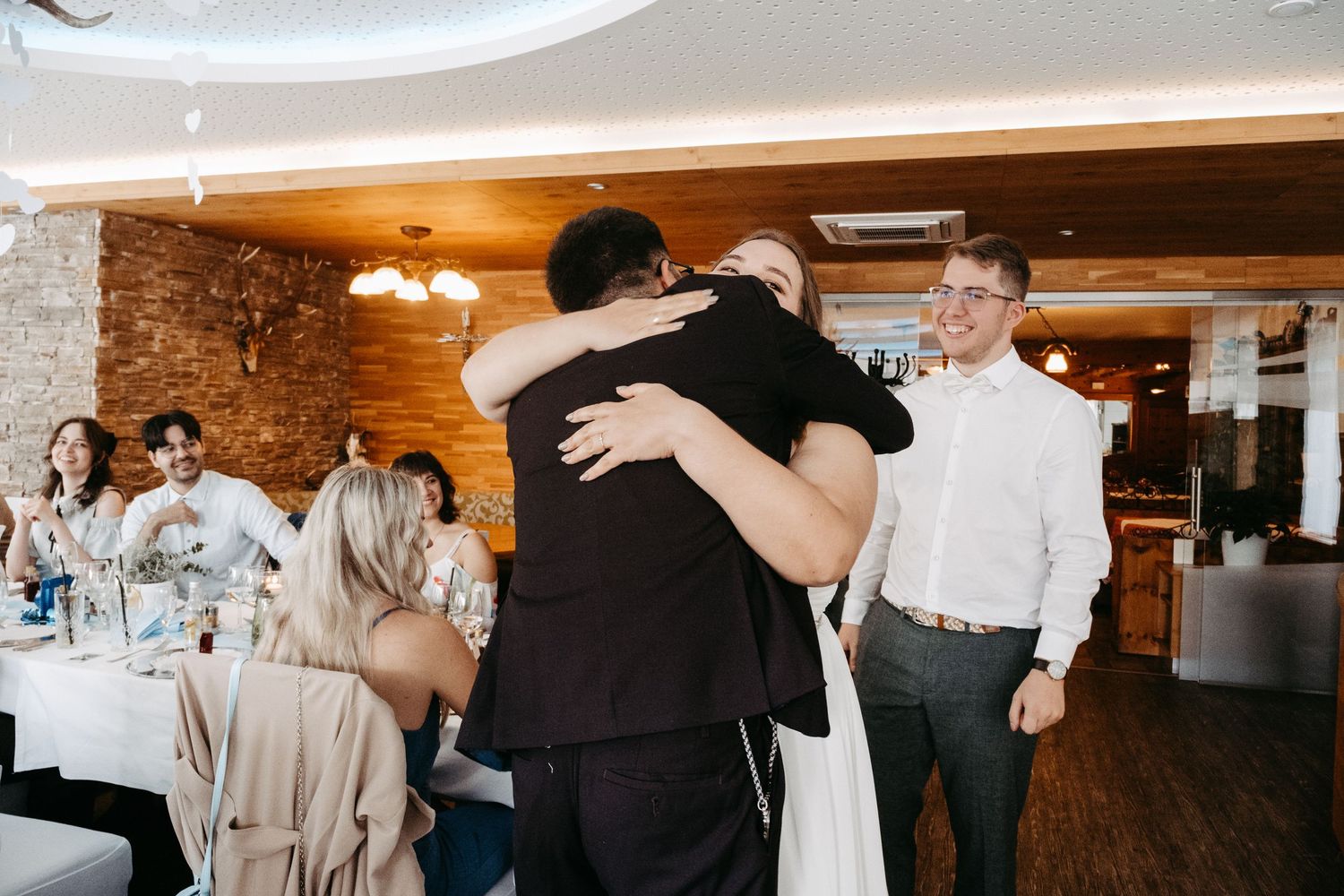 Two guests share a warm hug at a wedding reception while others smile nearby in an elegant venue.