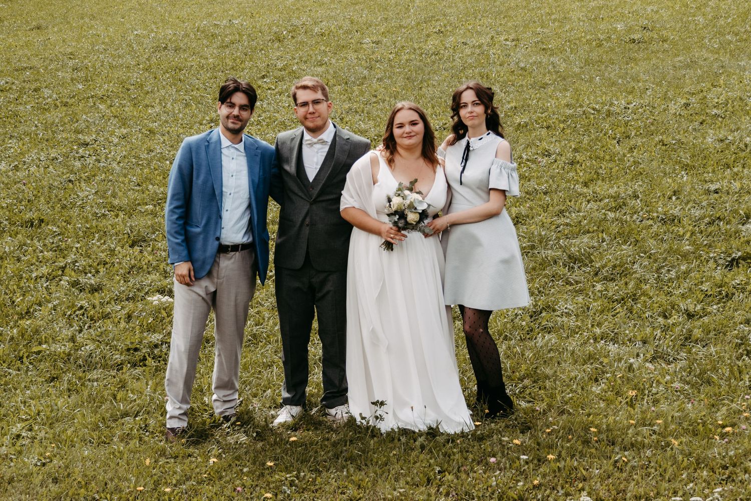 Wedding party of four posing on green grass field, bride in white gown holding bouquet with groom and two guests.