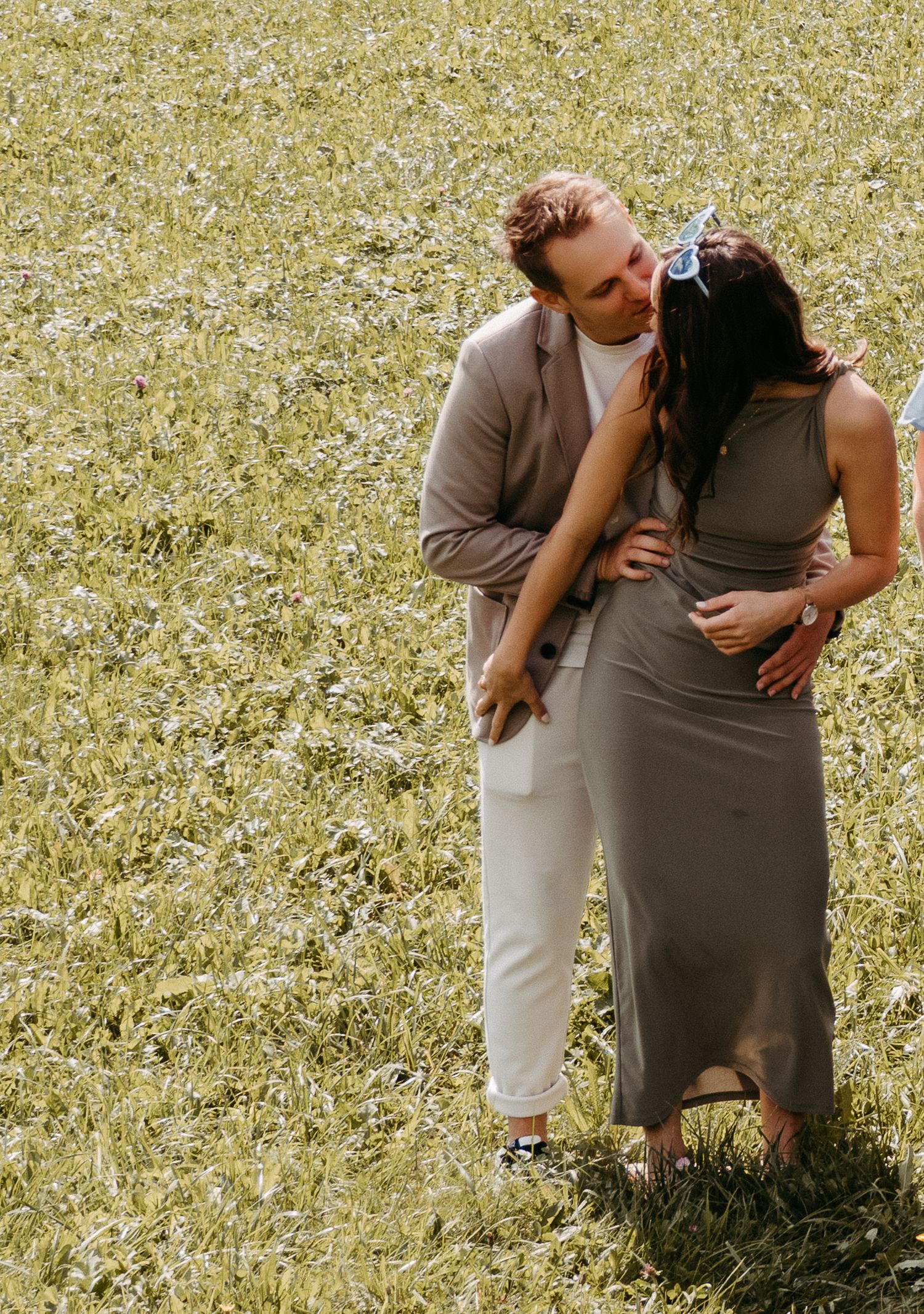 A couple shares a romantic kiss in a lush green field, man in tan blazer, woman in olive maxi dress.