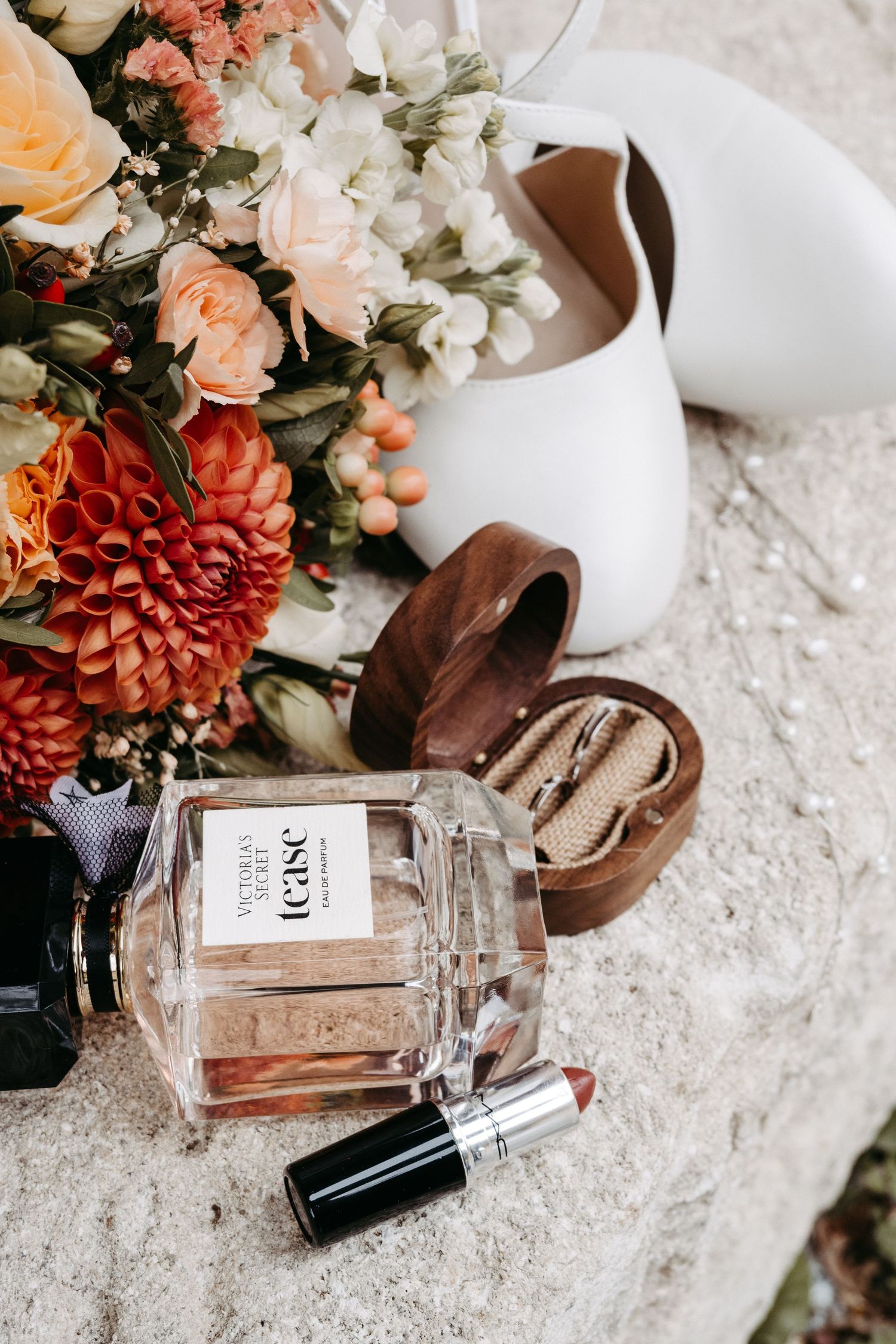 Bridal flat lay with Victoria's Secret Tease perfume, lipstick, ring box, white heels, and colorful floral bouquet.