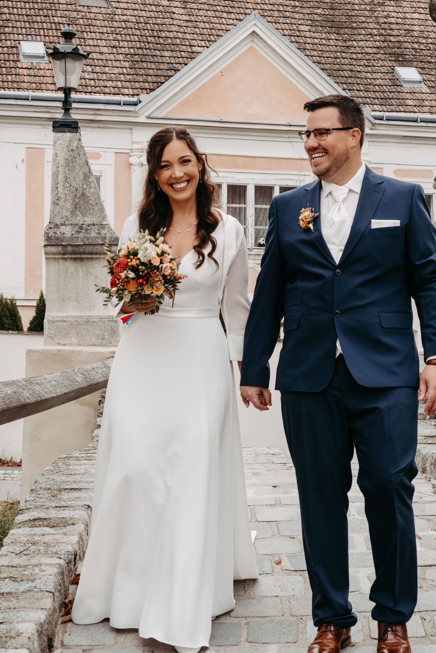 A couple in wedding attire walks along a stone wall pathway while holding hands and laughing together.