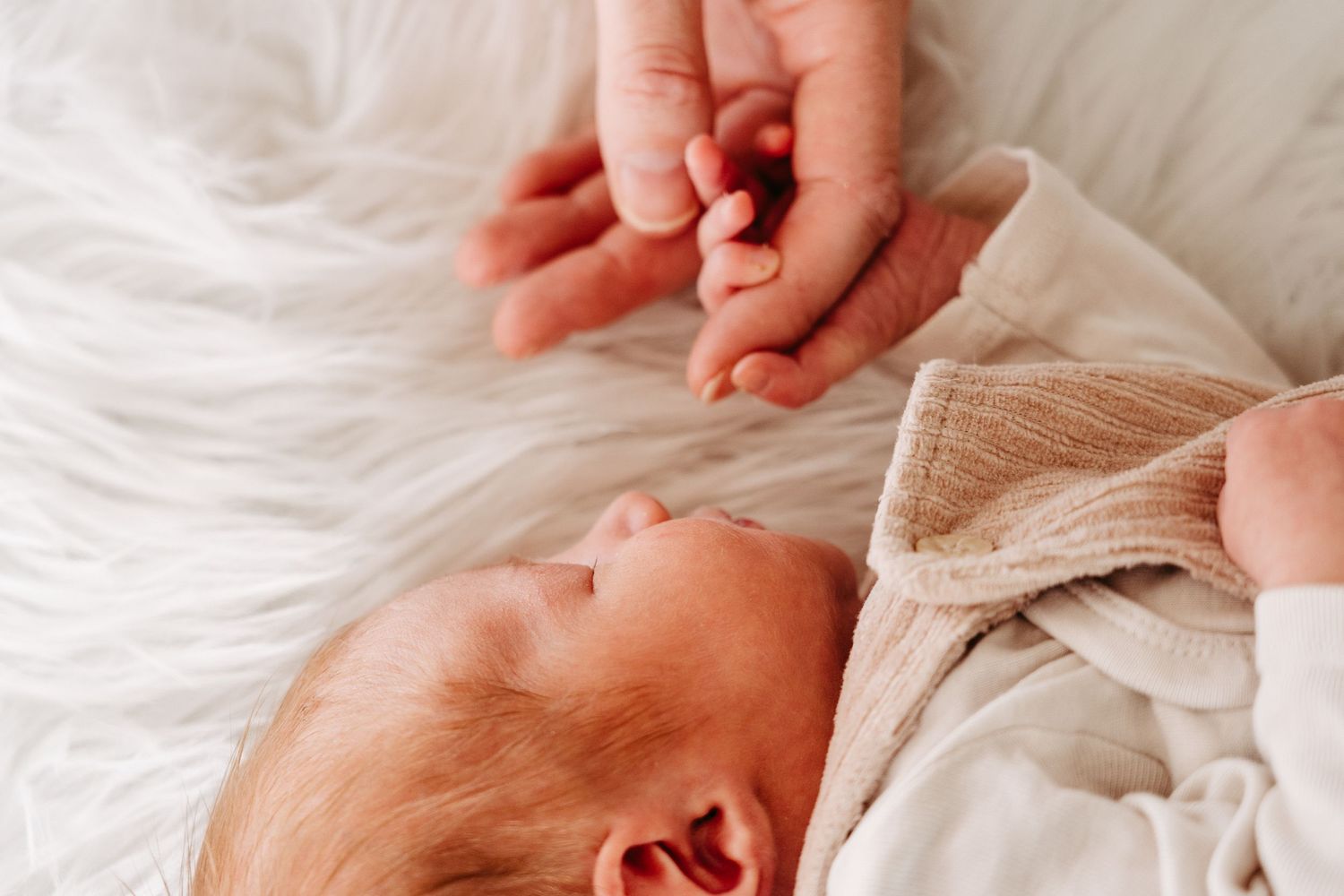 A close-up of a tiny hand grasping an adult's finger against white bedding.